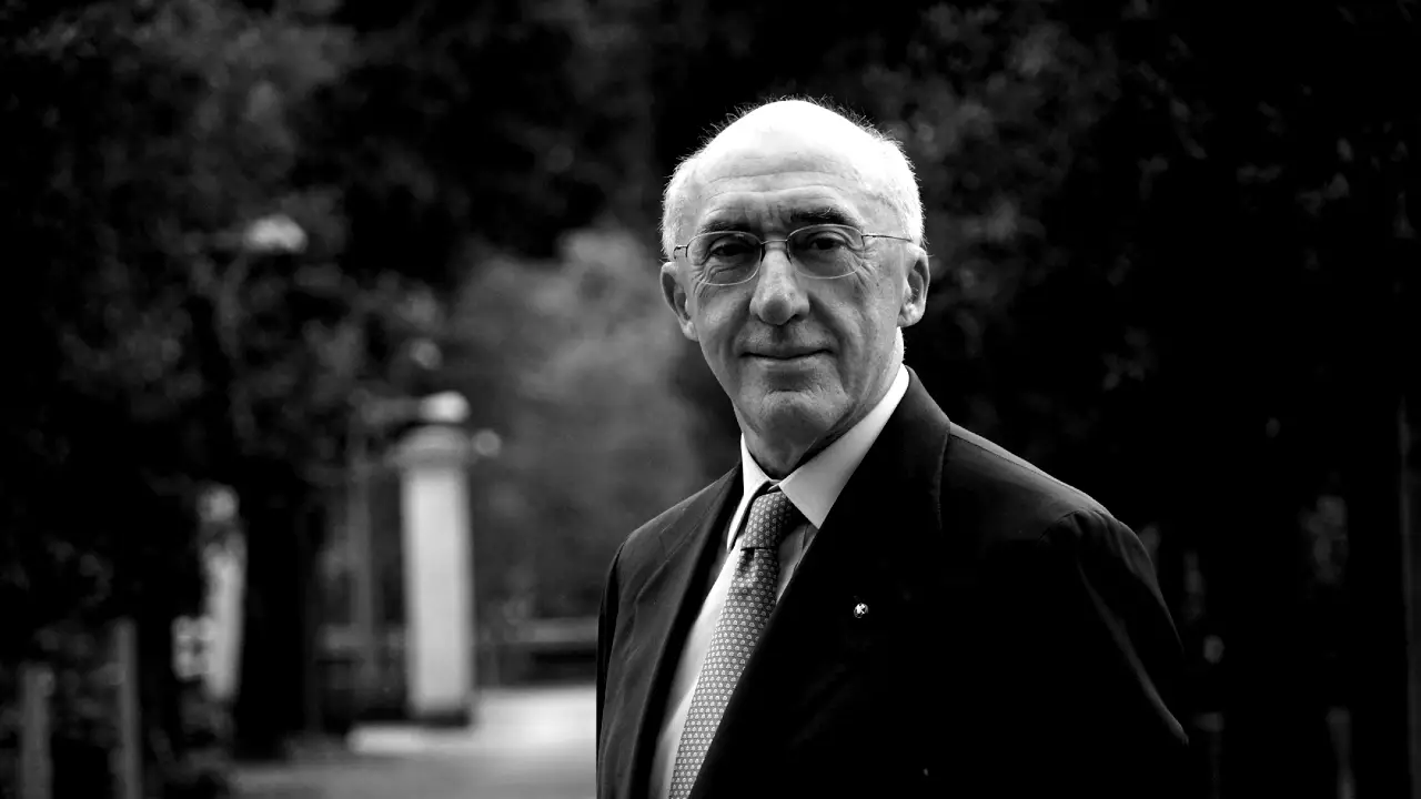 Un uomo anziano con gli occhiali e un completo si trova all'aperto, rivolto verso la macchina fotografica e con un leggero sorriso. L'immagine è in bianco e nero, con alberi e un sentiero sfocati sullo sfondo.