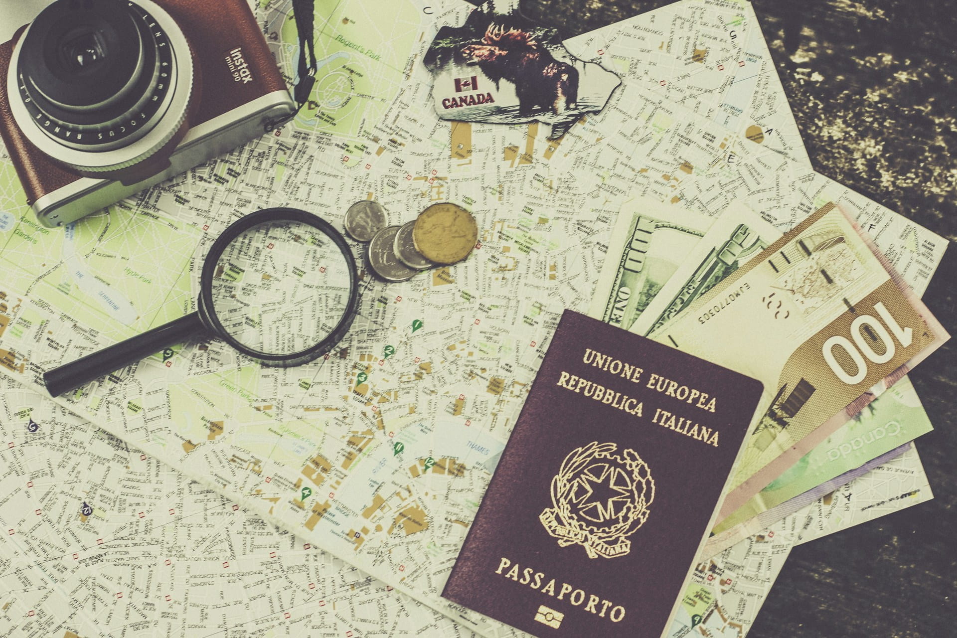 A camera, magnifying glass, coins, cash, Italy passport, and a Canada keychain are placed on a detailed city map, suggesting travel planning or preparation.