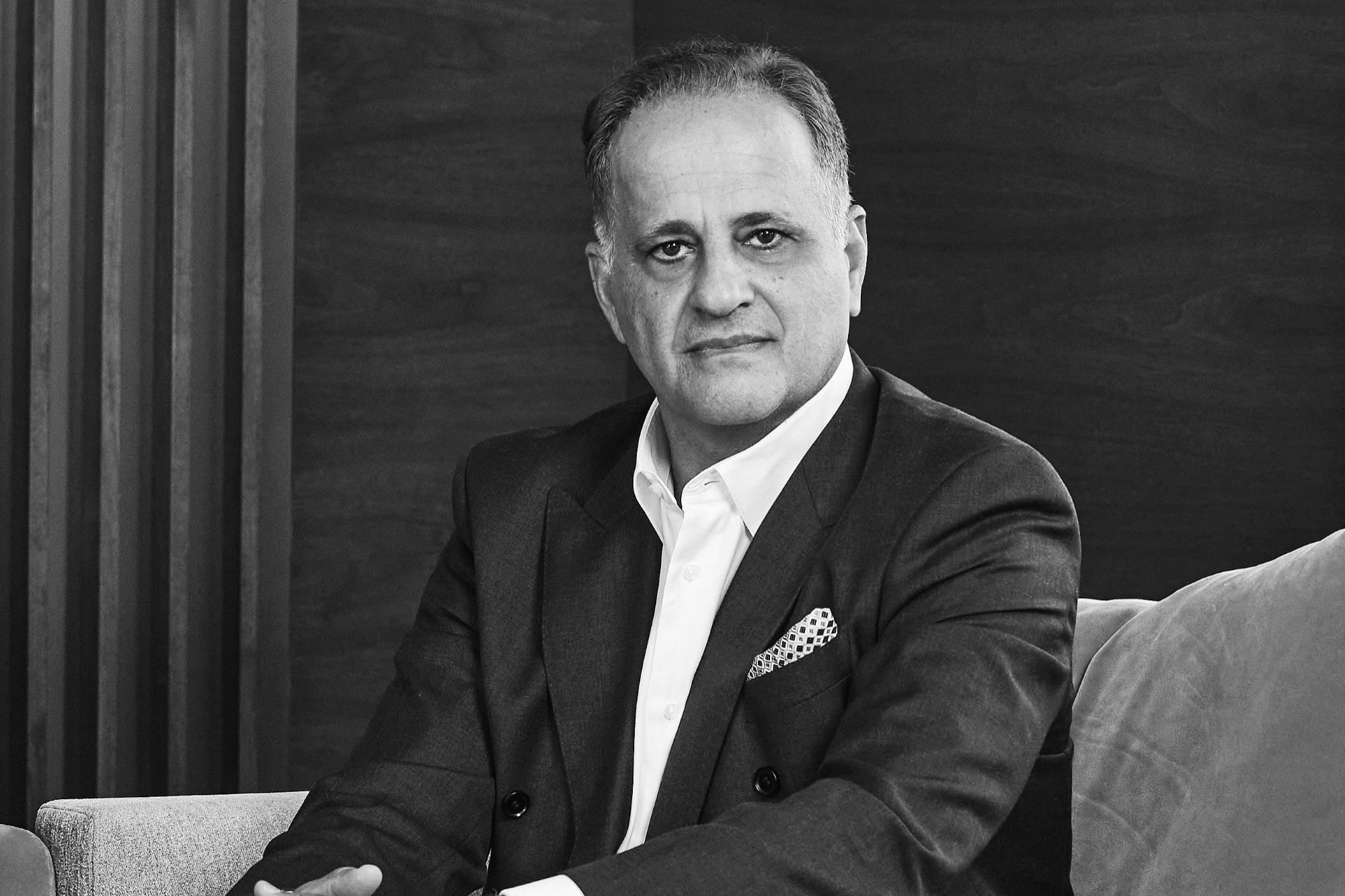 A man in a suit sits on a sofa, looking at the camera with a neutral expression. This black and white image, set against wooden panels, suggests someone ready to navigate venture capital, with a patterned pocket square accenting his jacket.