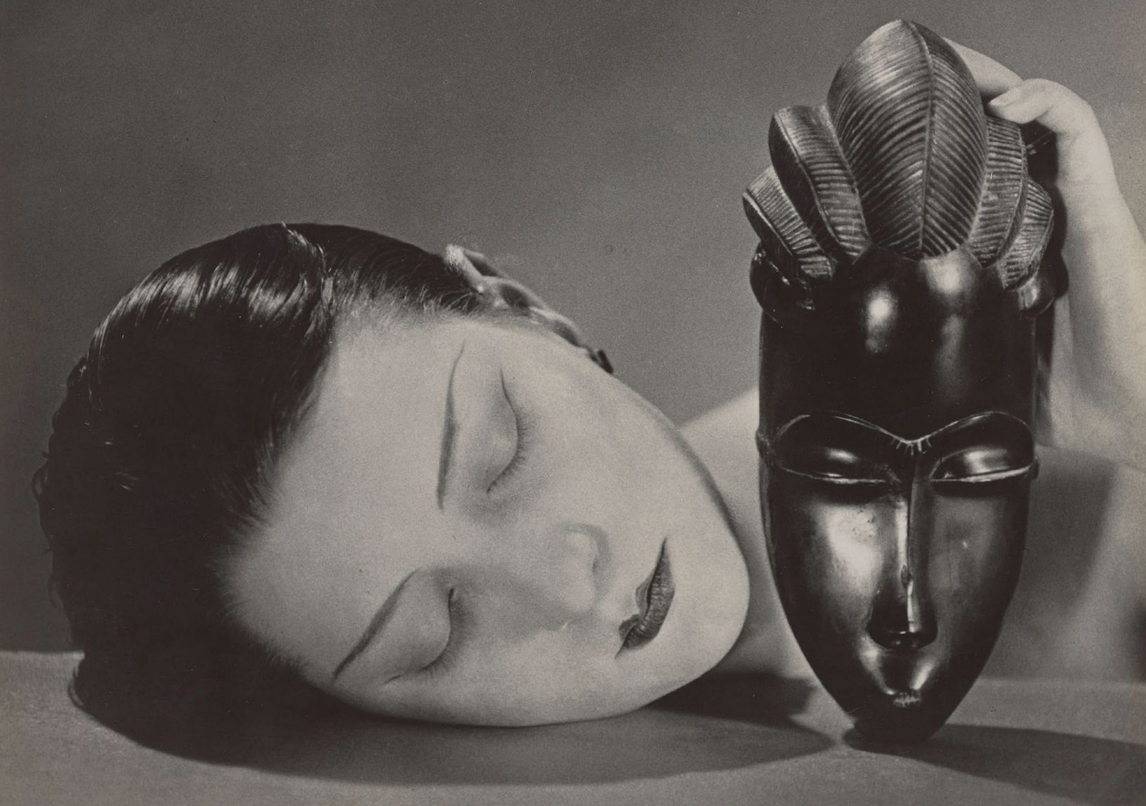 A woman with closed eyes rests her face on a surface next to an ornate, dark, sculpted mask that she holds upright. The composition highlights the contrast between her smooth skin and the mask’s intricate, glossy features.