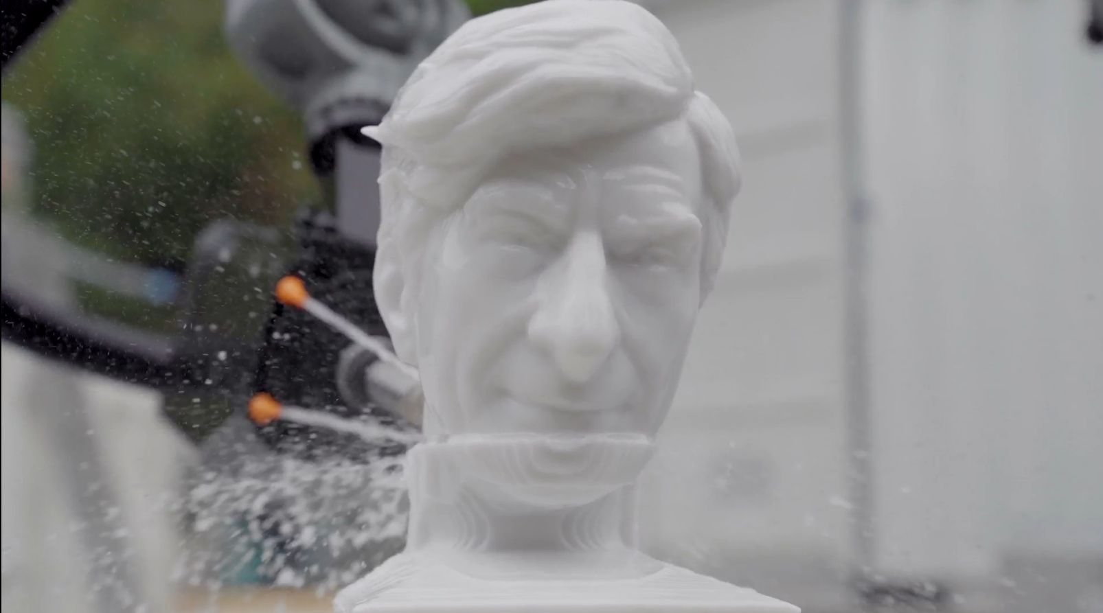 Un braccio robotico bianco spruzza acqua su un busto bianco stampato in 3D di un uomo con capelli e tratti del viso dettagliati, che ricordano lo stile di Maurizio Cattelan nel marmo, facendo schizzare acqua e particelle intorno alla scultura.
