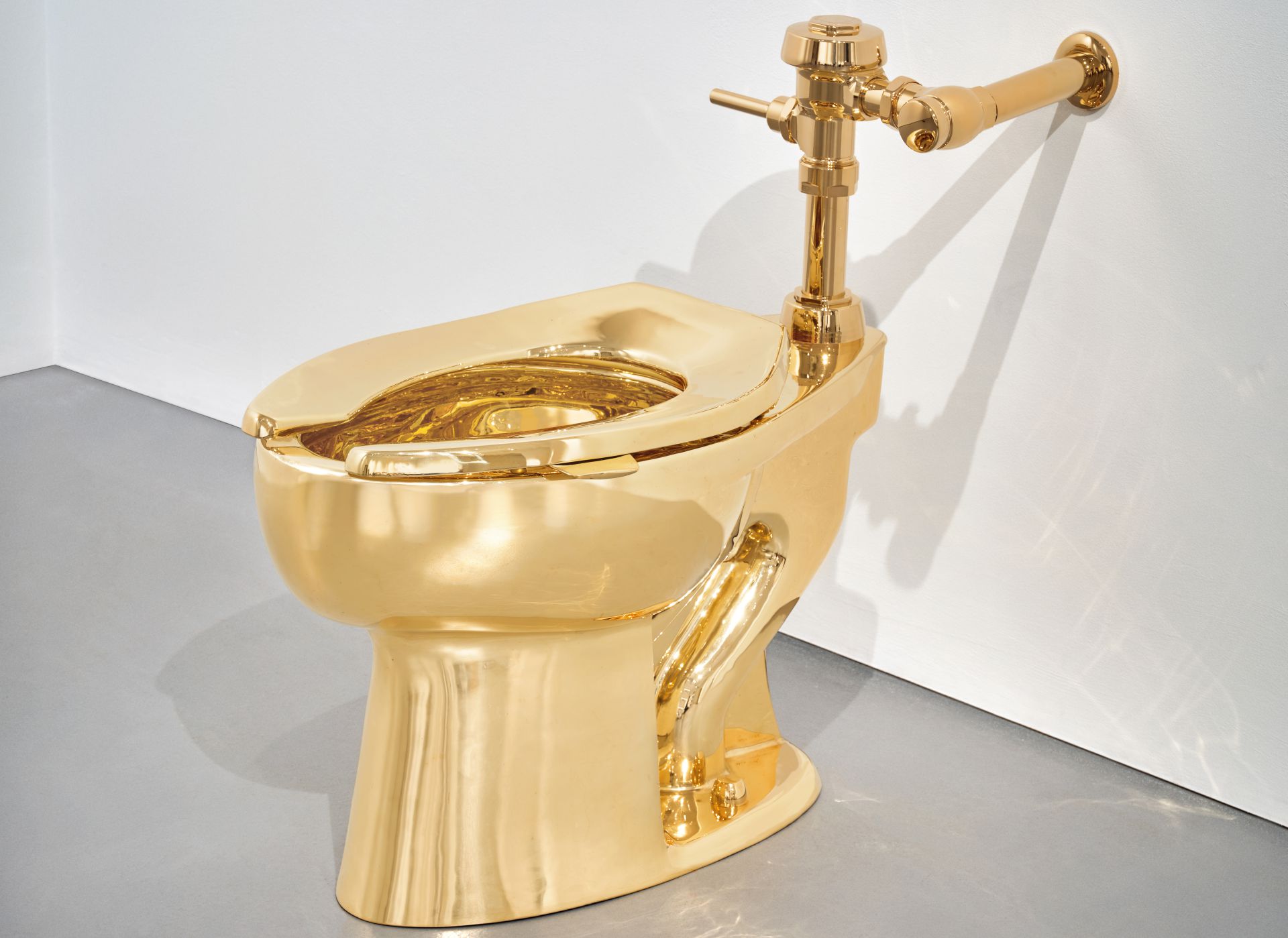 Una toilette interamente realizzata in oro lucido è installata in una stanza minimalista con pareti bianche e pavimento grigio. La toilette è dotata di impianti idraulici standard, anch'essi color oro.