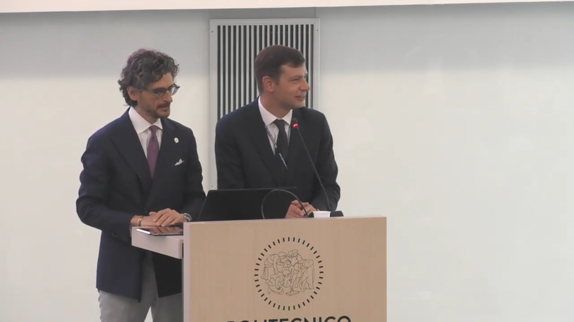 Due uomini in giacca e cravatta si trovano dietro un podio di legno con un computer portatile e un microfono e parlano a un pubblico. Il podio reca l'emblema e la scritta "POLITECNICO". Lo sfondo è semplice e di colore chiaro.