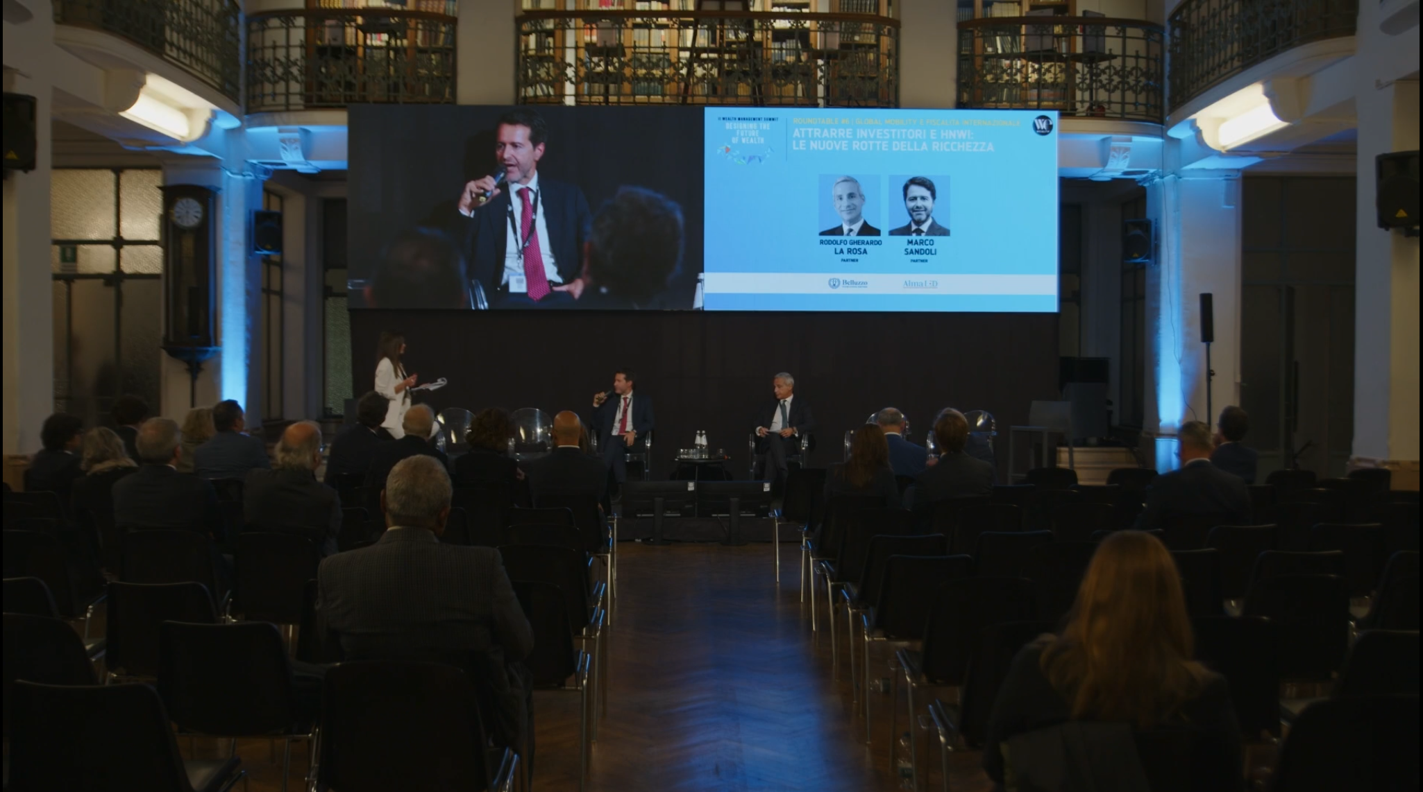 Una conferenza con un pubblico seduto di fronte a un palco dove tre relatori sono impegnati in una discussione. Un grande schermo in alto mostra una trasmissione video in diretta e i dettagli dell'evento. La sala è dotata di alti scaffali e di un'illuminazione calda.