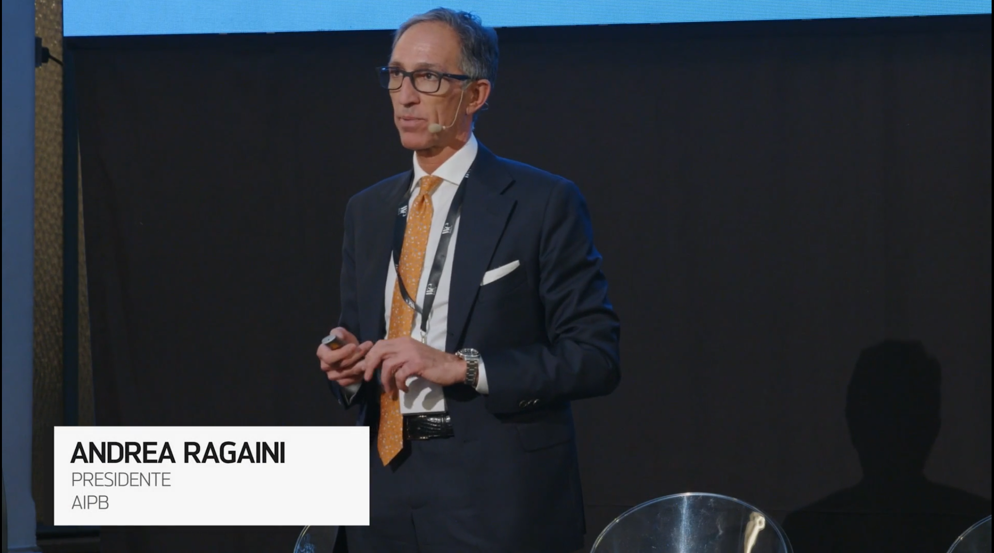 Un uomo in abito e cravatta arancione parla sul palco di una conferenza. Il testo sullo schermo recita: "Andrea Ragaini, Presidente, AIPB". Al suo fianco è visibile una sedia trasparente.