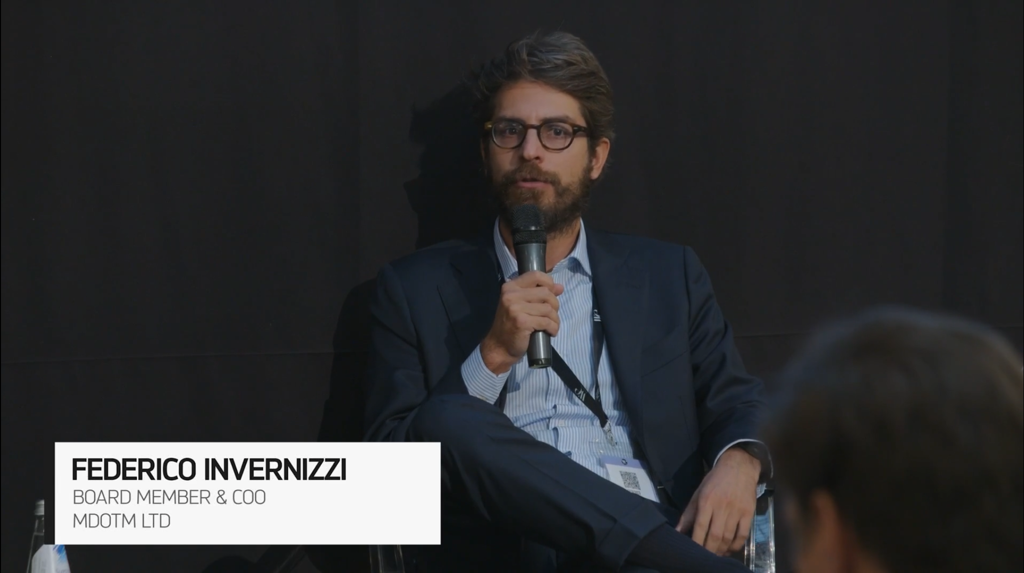 Un uomo in abito e occhiali parla al microfono seduto su un palco. Il testo in sovrimpressione recita: "Federico Invernizzi, Membro del Consiglio di Amministrazione e COO, Moditm Ltd.". Lo sfondo è scuro.