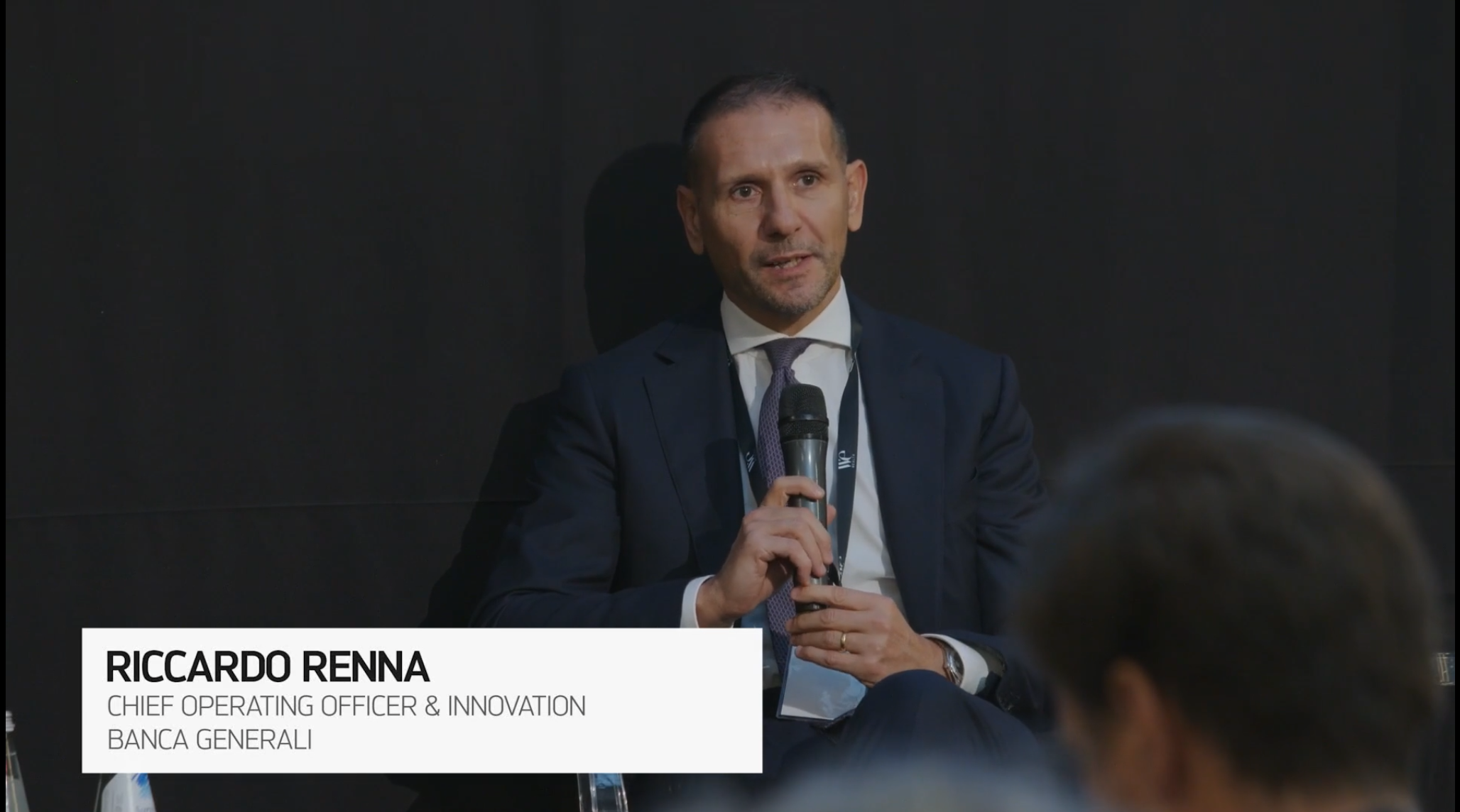 Un uomo in abito scuro siede e parla a un microfono durante un evento. La didascalia recita: "Riccardo Renna, Chief Operating Officer & Innovation, Banca Generali". Lo sfondo è scuro e sono visibili alcune teste del pubblico.