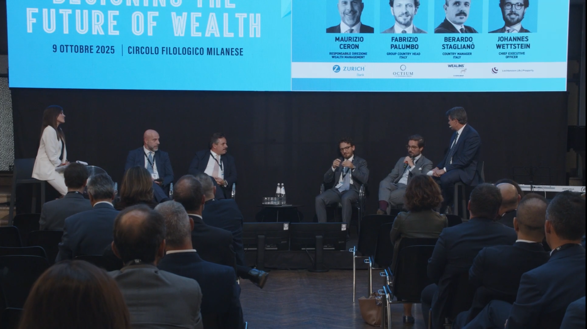 Un gruppo di sei relatori in abiti da lavoro siede sul palco di fronte a un pubblico, discutendo di gestione patrimoniale a un evento intitolato "Designing the Future of Wealth", con un grande schermo di presentazione blu alle loro spalle.