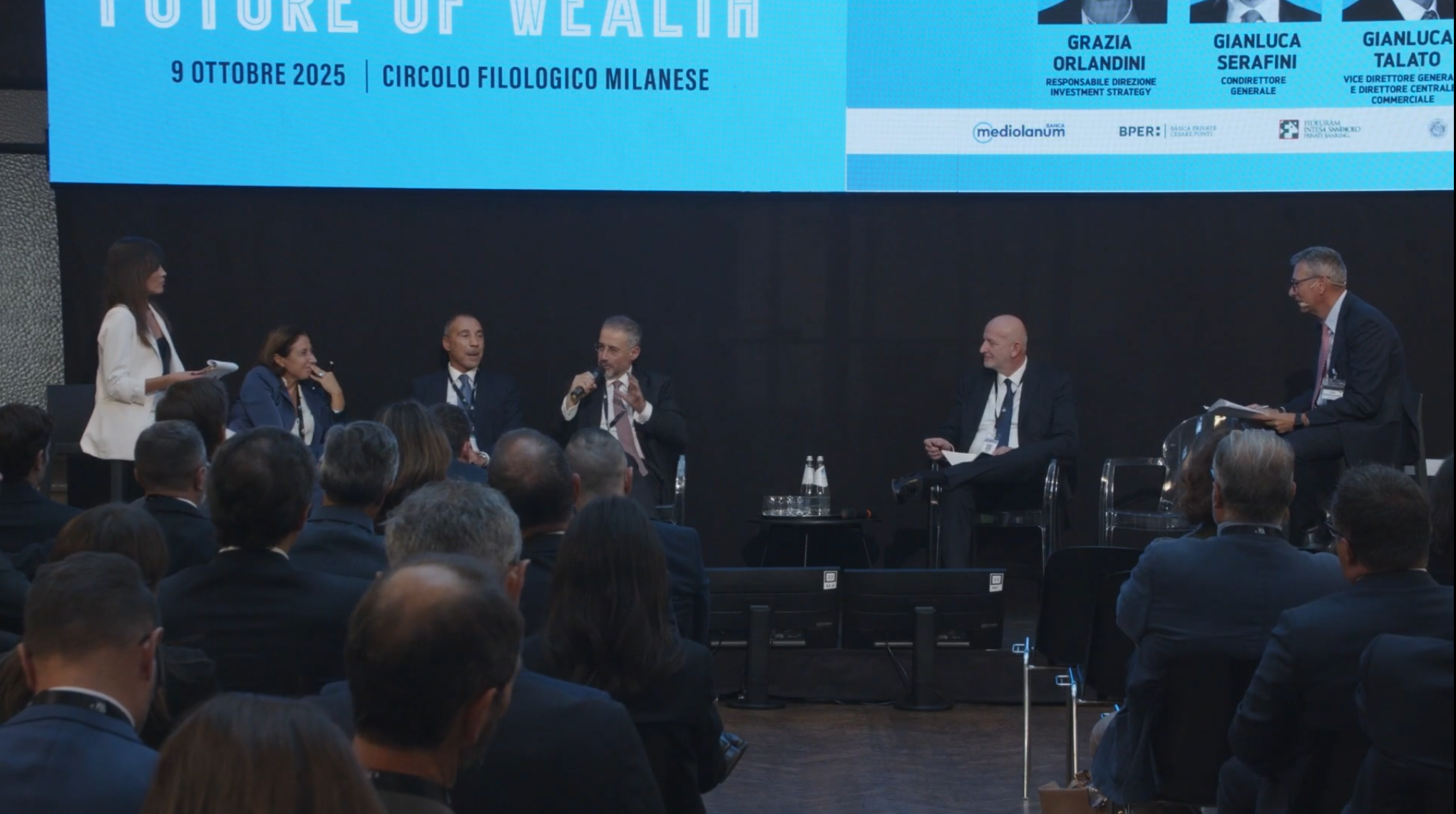 Un gruppo di cinque professionisti siede sul palco di una conferenza e parla al pubblico. Un grande schermo blu alle loro spalle mostra i dettagli dell'evento, tra cui "Future of Wealth", la data e i nomi dei relatori.