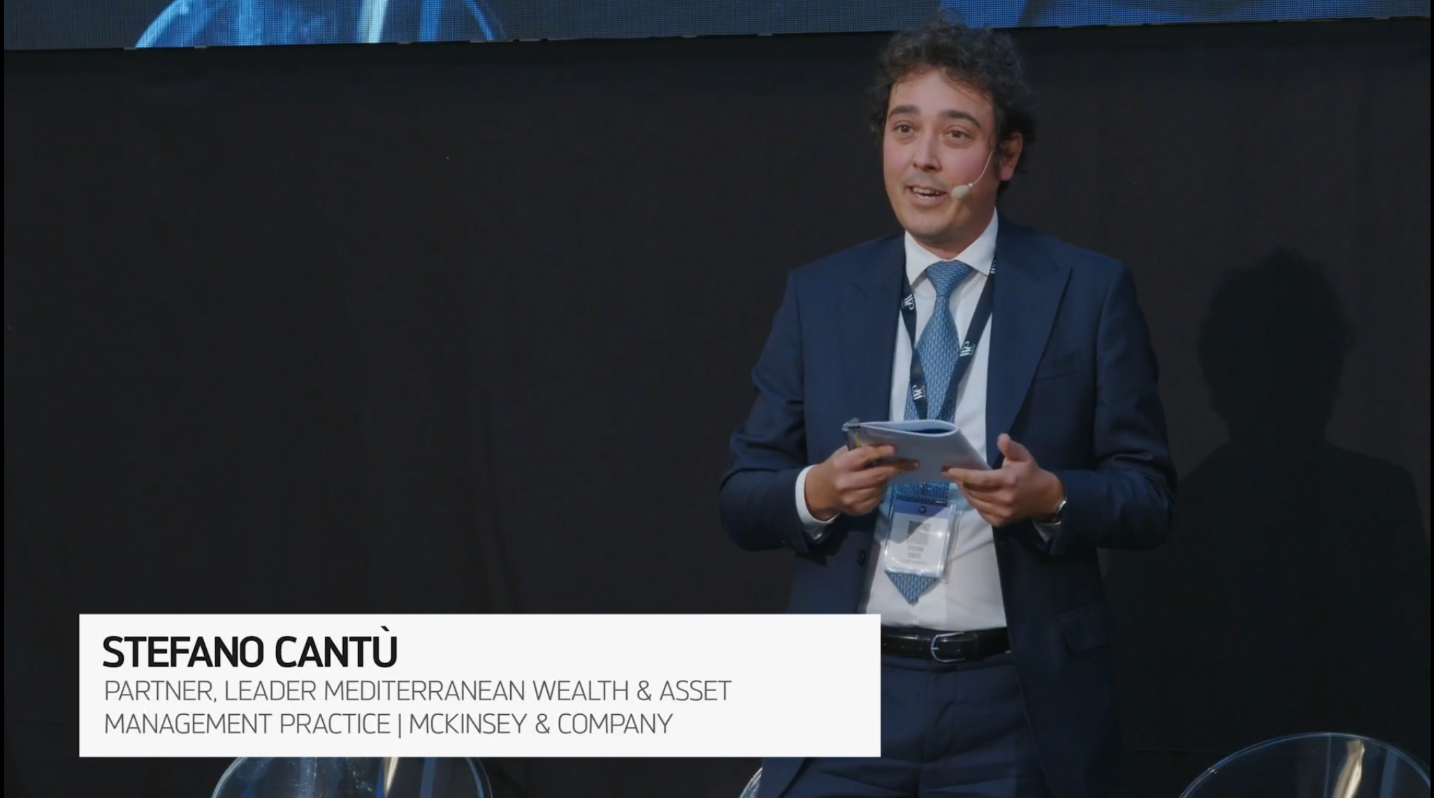 Un uomo in abito blu sta sul palco con dei documenti in mano e parla a un evento. Un riquadro di testo lo identifica come Stefano Cantù, Partner e Leader della Mediterranean Wealth & Asset Management Practice di McKinsey & Company.