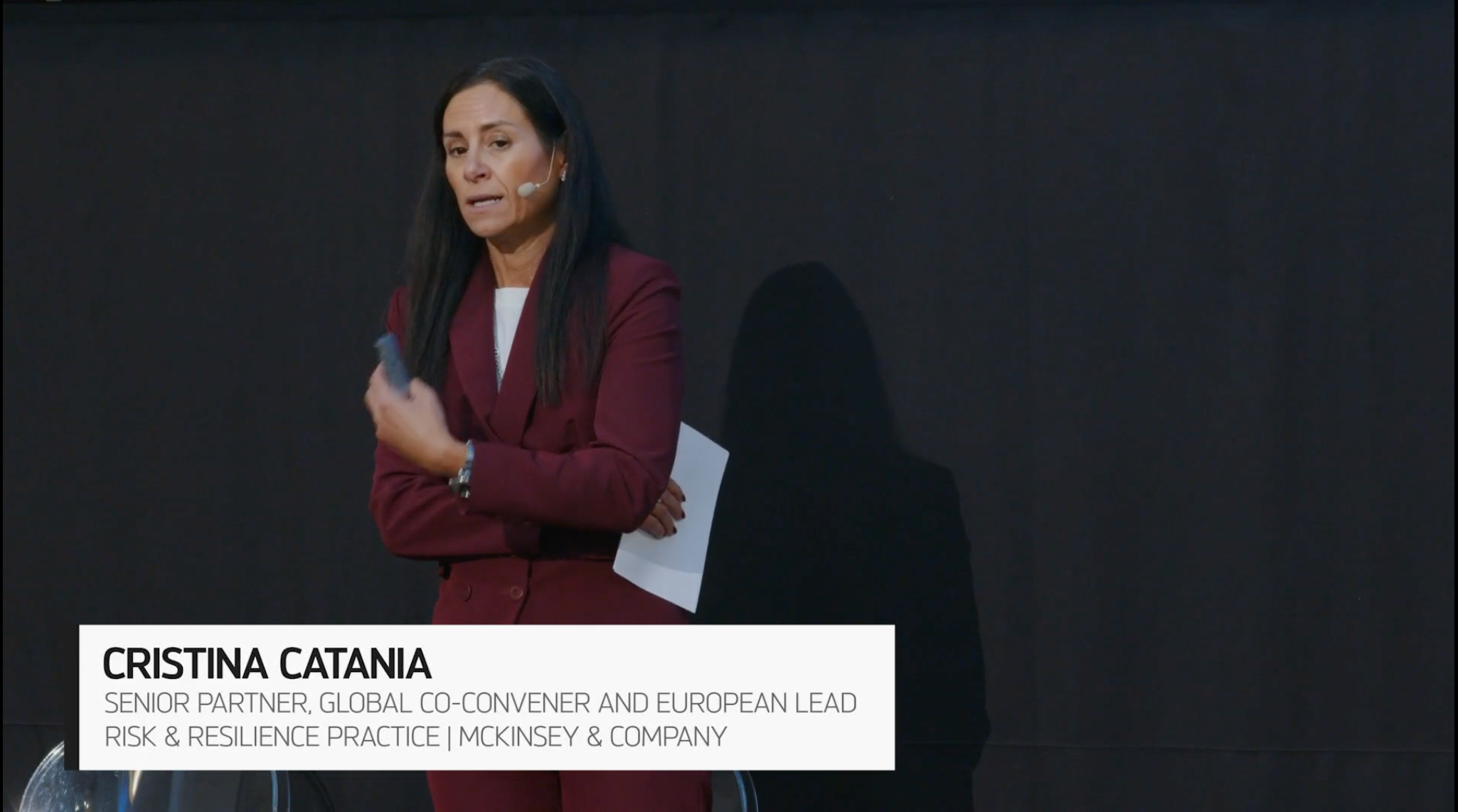 Una donna in abito marrone si staglia su uno sfondo scuro, tenendo in mano un foglio e gesticolando con una mano. Un riquadro di testo la identifica come Cristina Catania, Senior Partner di McKinsey & Company.