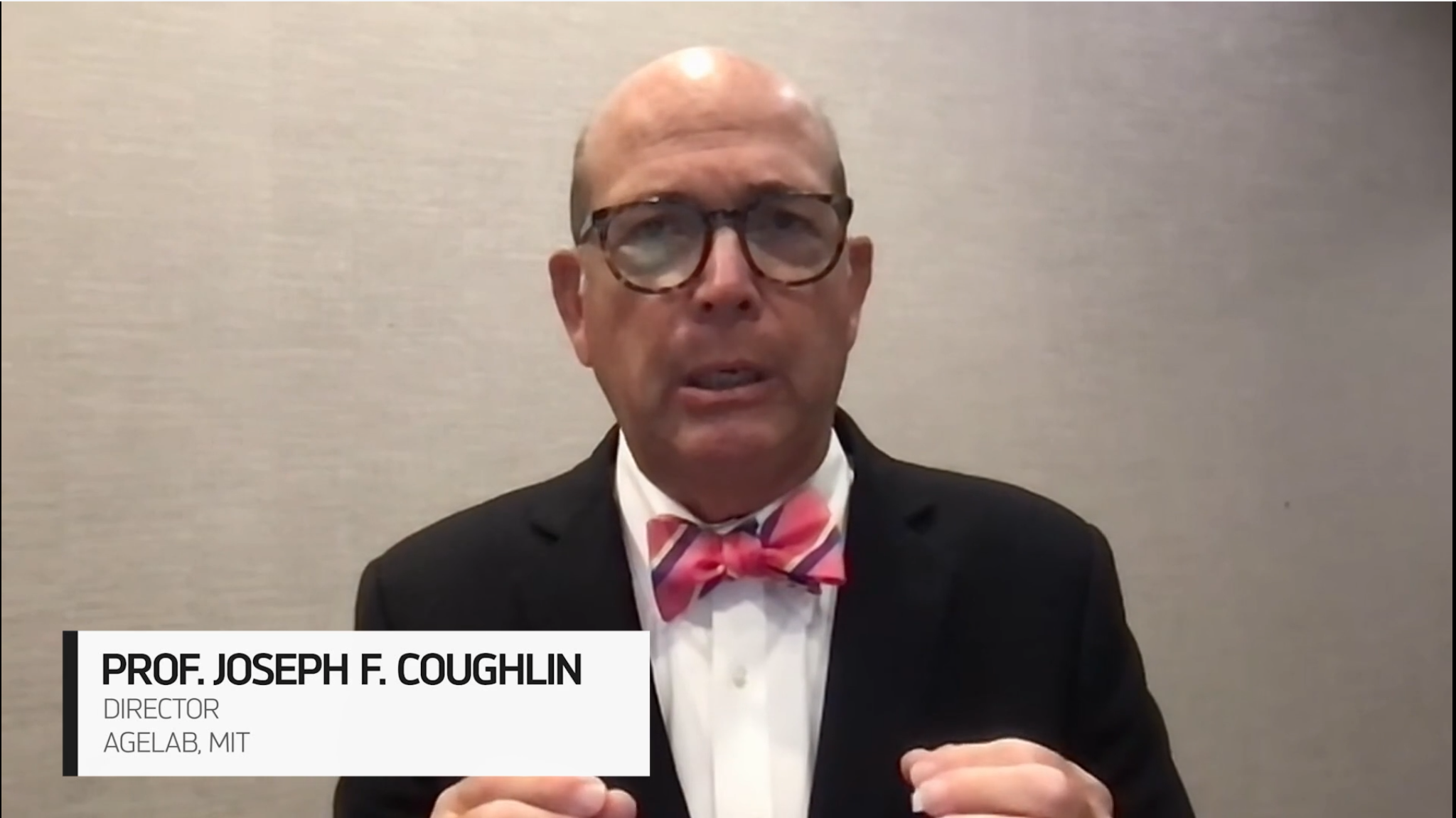 Un uomo con occhiali, abito scuro e papillon rosa parla davanti a uno sfondo semplice, discutendo di longevita demografica. Un'etichetta in basso recita: "Prof. Joseph F. Coughlin, Direttore, AgeLab, MIT.