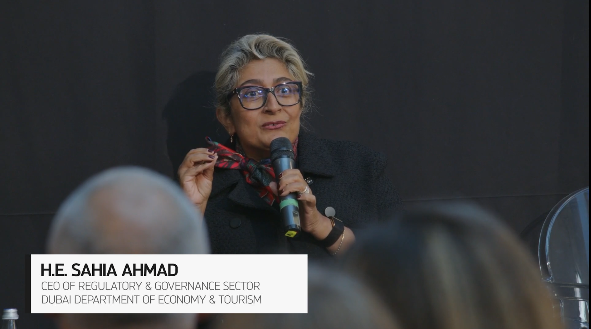 Una donna con capelli corti grigi e occhiali parla a un microfono durante un evento. Indossa una giacca scura e una sciarpa rossa a fantasia. Un cartellino sullo schermo recita: S.E. Sahia Ahmad, CEO del settore Regolamentazione e Governance, Dipartimento dell'Economia e del Turismo di Dubai.