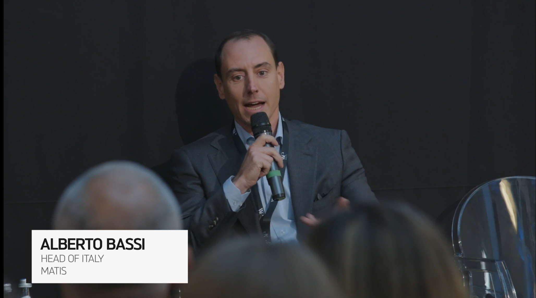 Un uomo in giacca e cravatta parla al microfono durante un evento. In primo piano sono visibili le teste sfocate dei membri del pubblico. Un riquadro di testo recita "ALBERTO BASSI, Responsabile Italia, MATIS.