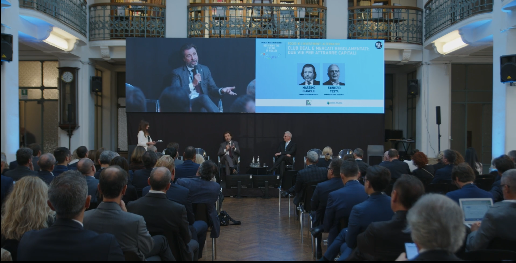 Una conferenza con molti partecipanti in giacca e cravatta siede di fronte a un palco. Tre relatori parlano sul palco, mentre alle loro spalle un grande schermo mostra i nomi dei due relatori, le foto e il titolo dell'evento in italiano. La sede è dotata di librerie e ringhiere.