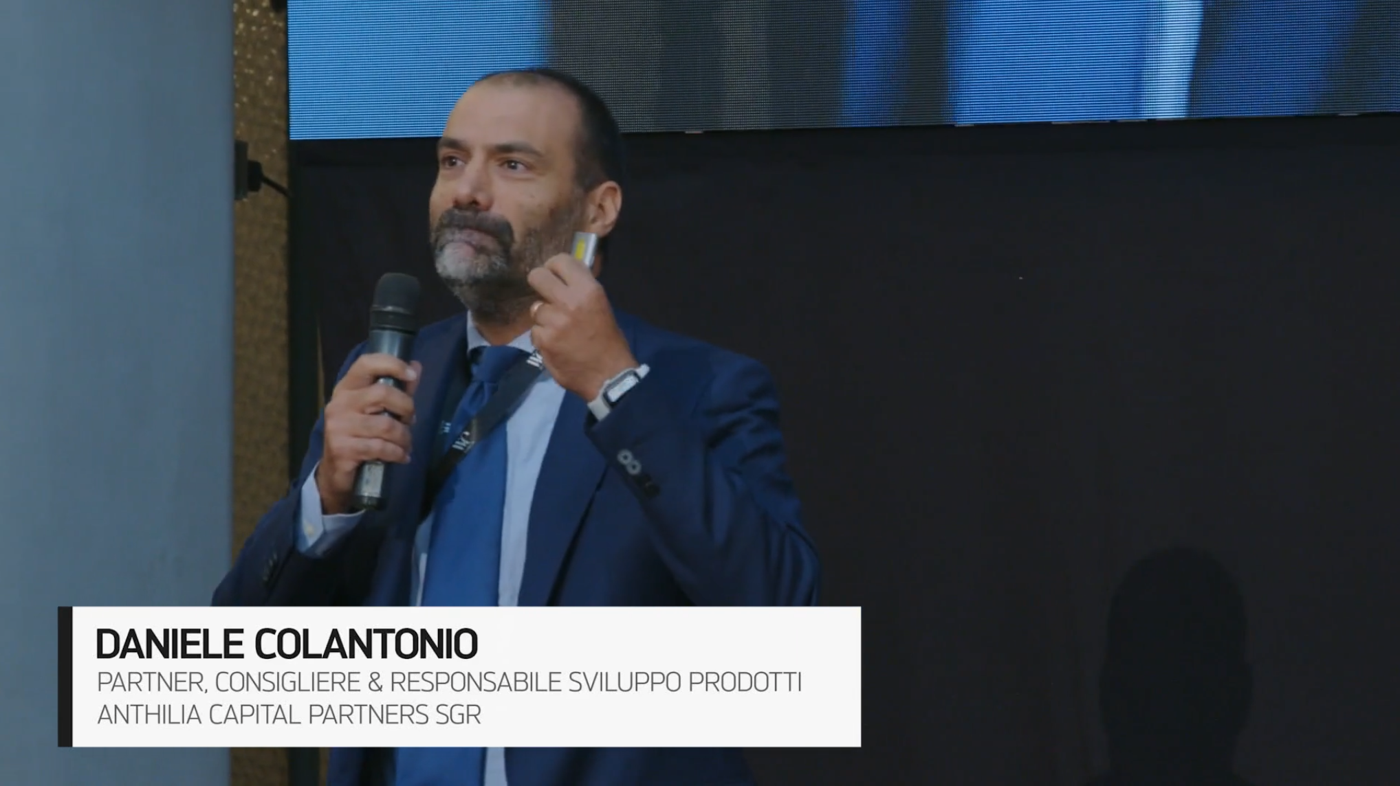 Un uomo in abito blu con un microfono in mano parla sul palco. Una casella di testo nell'angolo in basso a sinistra mostra il suo nome, Daniele Colantonio, il suo titolo professionale e i dettagli della sua azienda.
