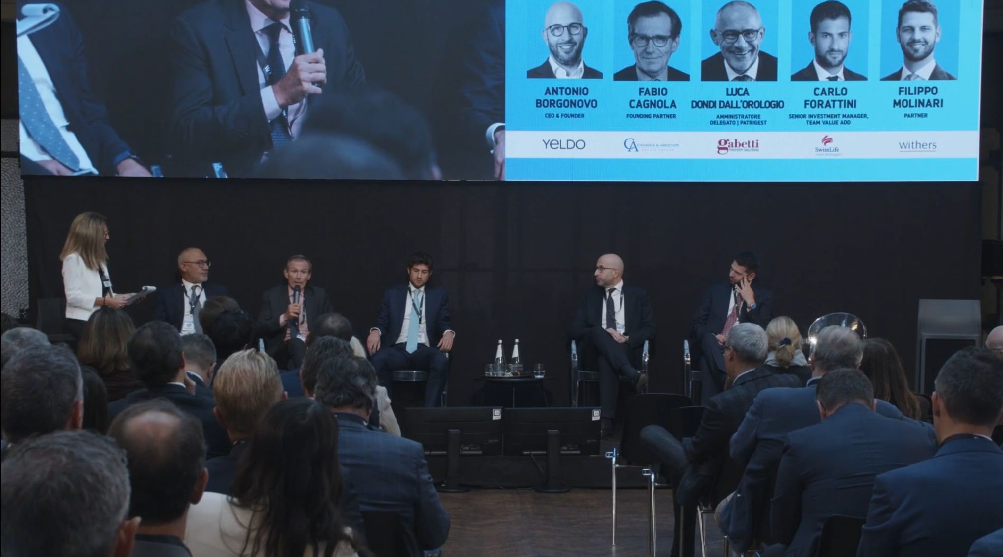 Un gruppo di sei uomini in giacca e cravatta siede sul palco durante una conferenza, con una moderatrice donna in piedi. Il pubblico è seduto. Un grande schermo in alto mostra le foto, i nomi e i titoli dei relatori.