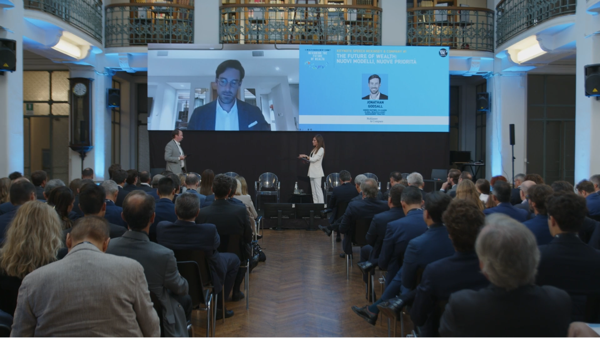 Un pubblico numeroso siede in una sala conferenze di fronte a un palco, dove parlano un uomo e una donna. Un grande schermo alle loro spalle mostra una videochiamata con un uomo e i dettagli dell'evento su "Il futuro della salute" e il suo impatto sulla ricchezza futura.