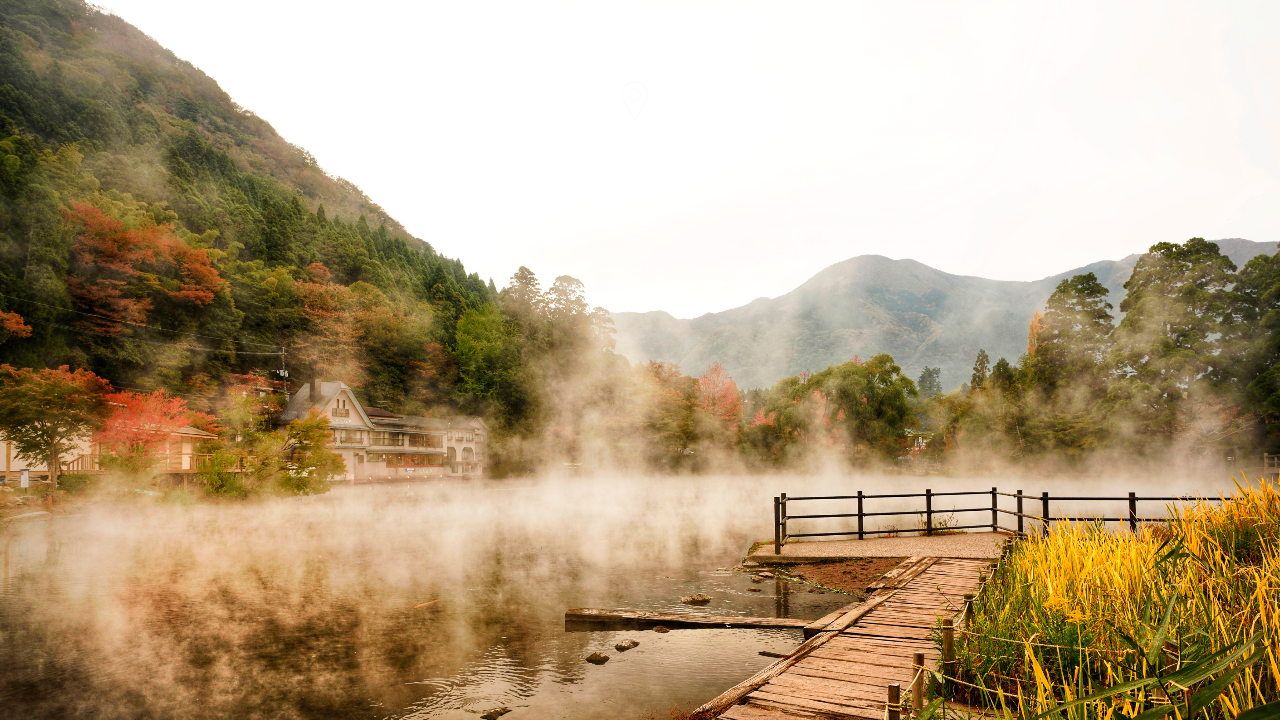 Una scena nebbiosa in riva al lago con un molo di legno, circondato da erbe gialle, alberi con fogliame autunnale e montagne sullo sfondo. Gli edifici tradizionali sono parzialmente visibili attraverso la nebbia.