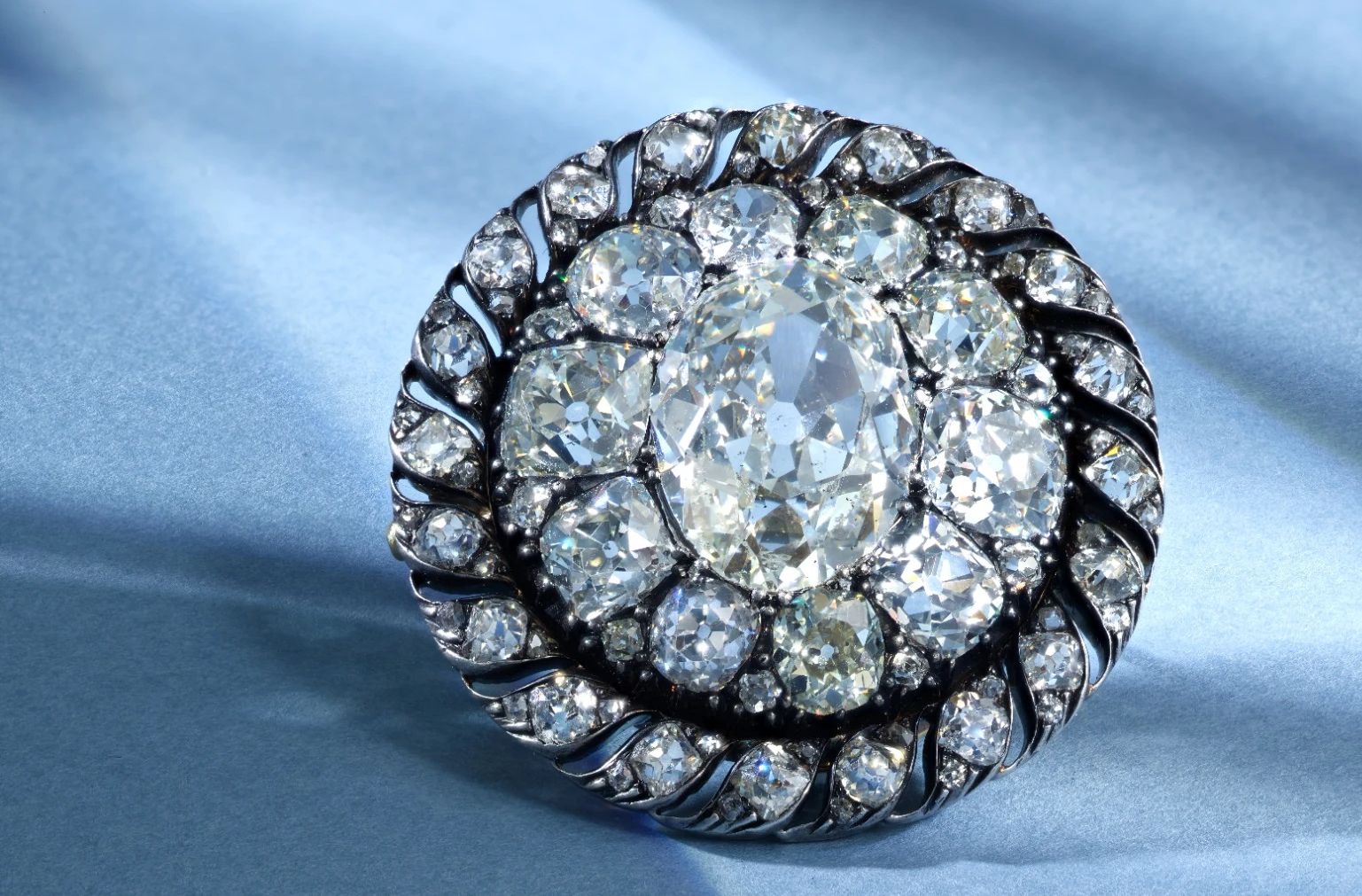 Spilla vintage con un grande diamante centrale di taglio ovale, circondato da due file di diamanti rotondi più piccoli, incastonati in un'ornamentale montatura di metallo in stile antico su uno sfondo azzurro.