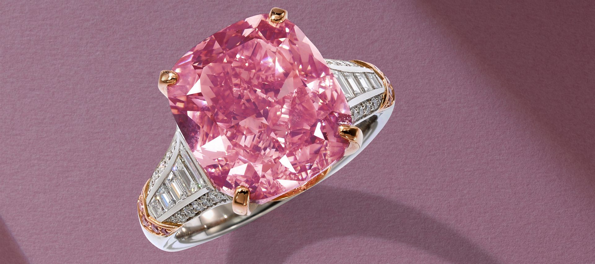 Un anello in argento con al centro un grande diamante rosa taglio cuscino, incastonato in un castone d'oro e con bande laterali di diamanti bianchi, è elegantemente esposto su una superficie color malva che ricorda le migliori vetrine di Sotheby.