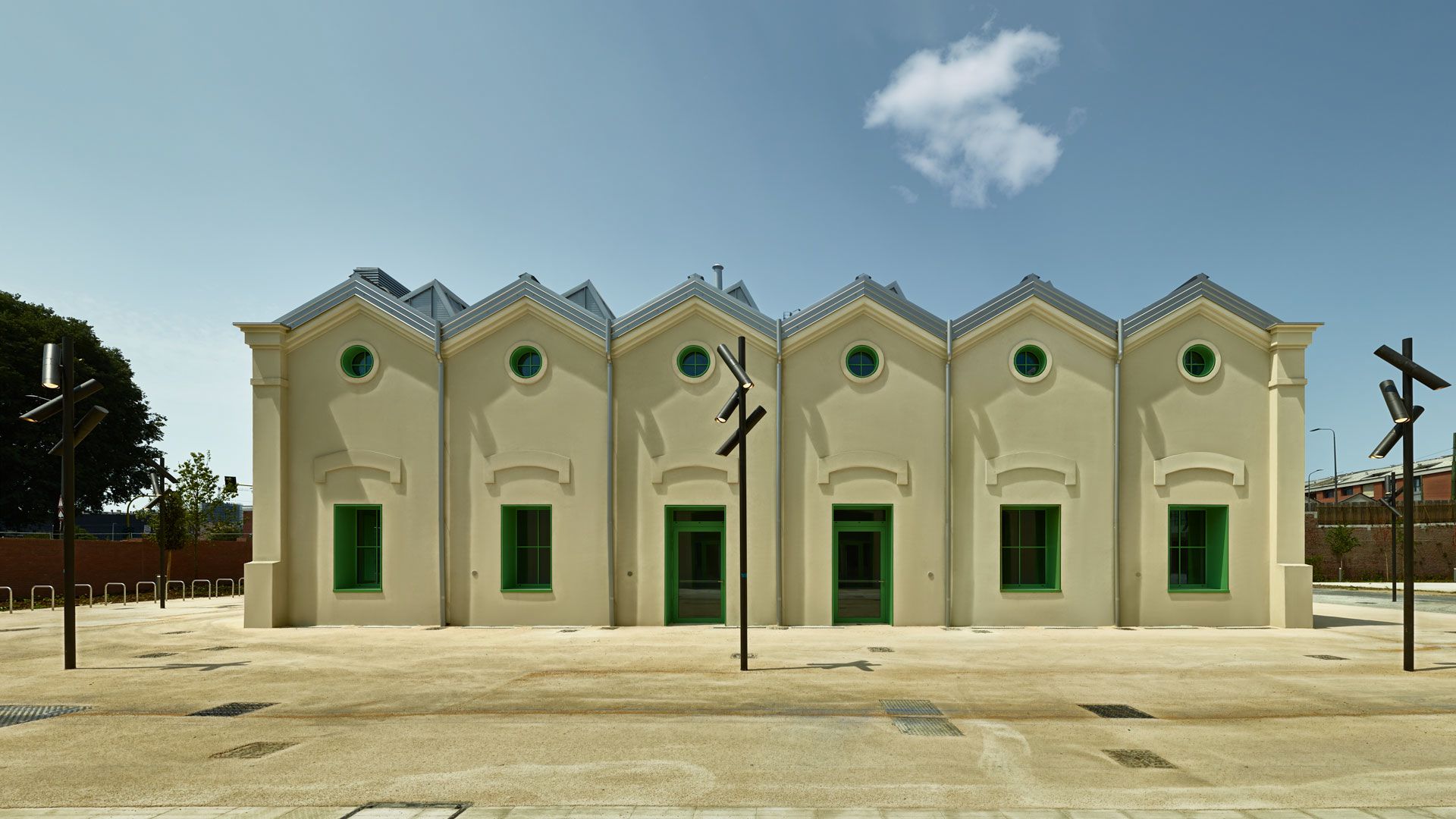 Un edificio beige simmetrico con cinque tetti a spiovente, finestre e porte con cornice verde e finestre rotonde sopra ogni ingresso. L'edificio si trova su un'ampia area pavimentata vuota, sotto un cielo azzurro con poche nuvole.