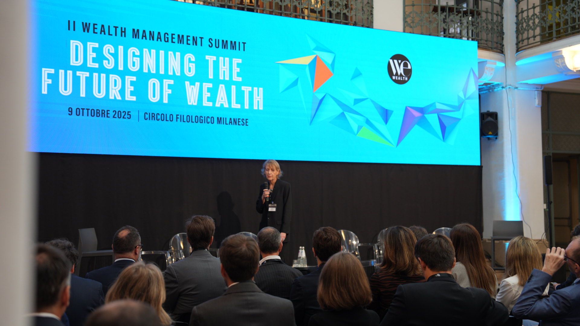 Fabienne Mailfait sul palco parla al pubblico del Wealth Management Summit 2025. Alle sue spalle, un grande schermo mostra il titolo dell'evento "Designing the Future of Wealth", oltre alla data e alla sede di Milano.