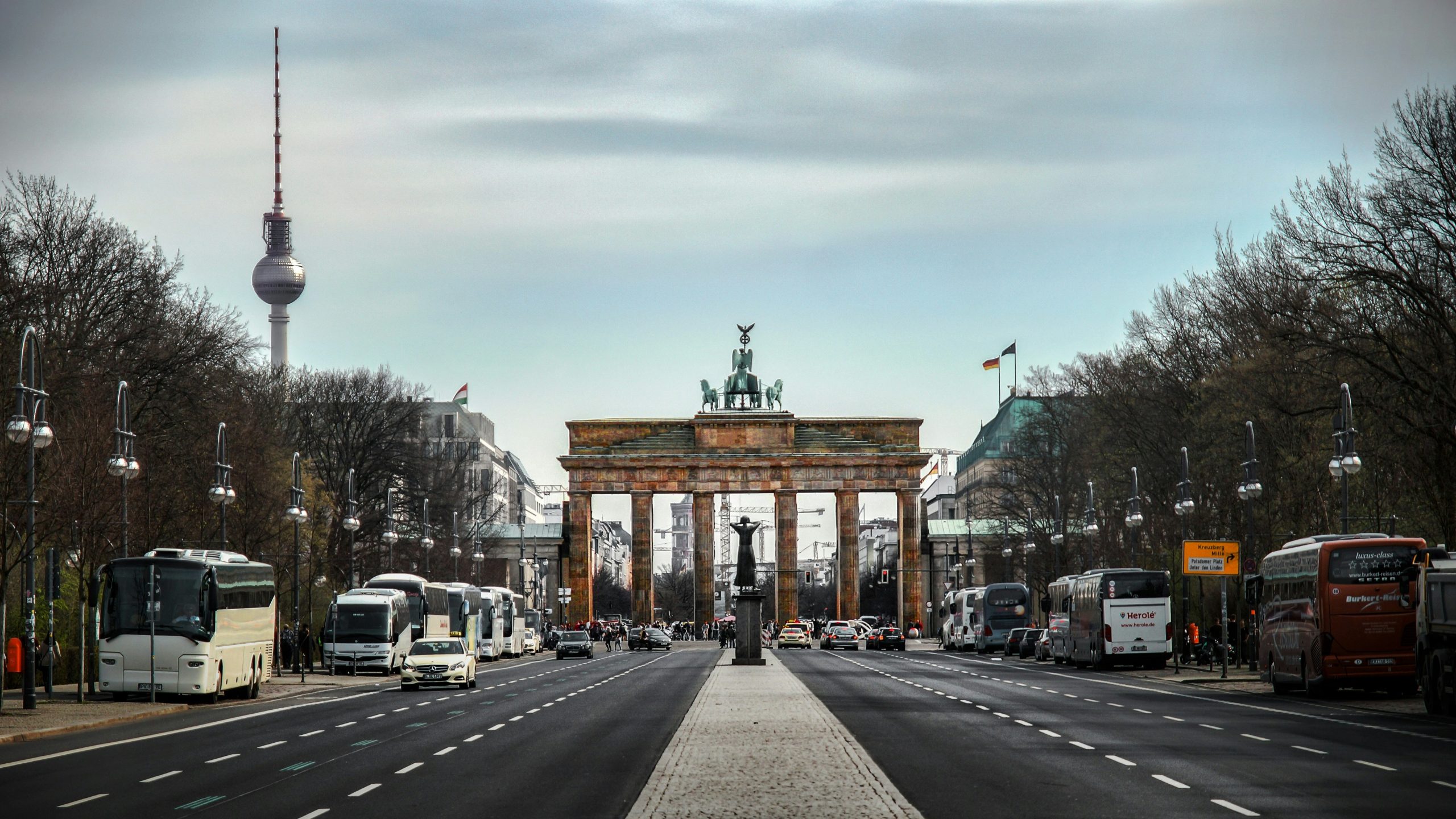 Ampio viale che conduce alla Porta di Brandeburgo di Berlino, con auto e autobus su entrambi i lati, alberi lungo la strada e la torre televisiva Fernsehturm visibile sullo sfondo sotto un cielo nuvoloso.