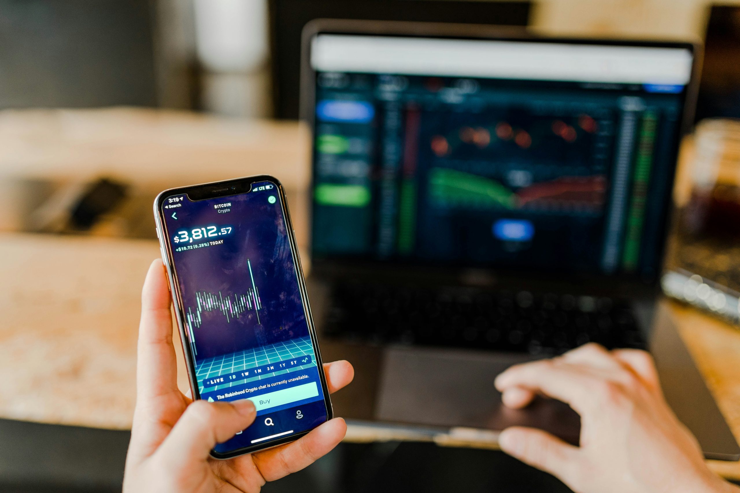 Una persona tiene in mano uno smartphone che visualizza un grafico del mercato azionario, con i grafici di Trade Republic Reddito Fisso sul proprio laptop sullo sfondo. L'ambiente sembra essere uno spazio di lavoro o un ufficio domestico.