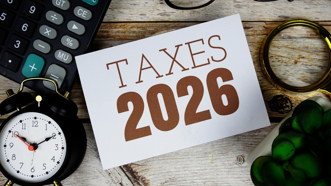Un cartoncino bianco con la scritta "TAXES 2026" in grassetto marrone si trova su una superficie di legno con una calcolatrice. Il riferimento va alle novità della manovra 2026 su casa e famiglia.
