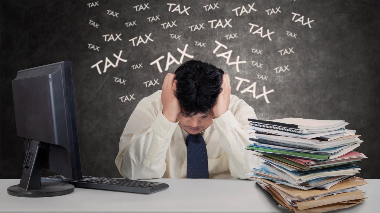 Un uomo stressato siede alla scrivania con la testa tra le mani, circondato da scartoffie e da un computer, mentre sulla lavagna alle sue spalle compare ripetutamente la parola "TAX". Il riferimento va alla stretta dell’Agenzia delle Entrate sui carried interest.