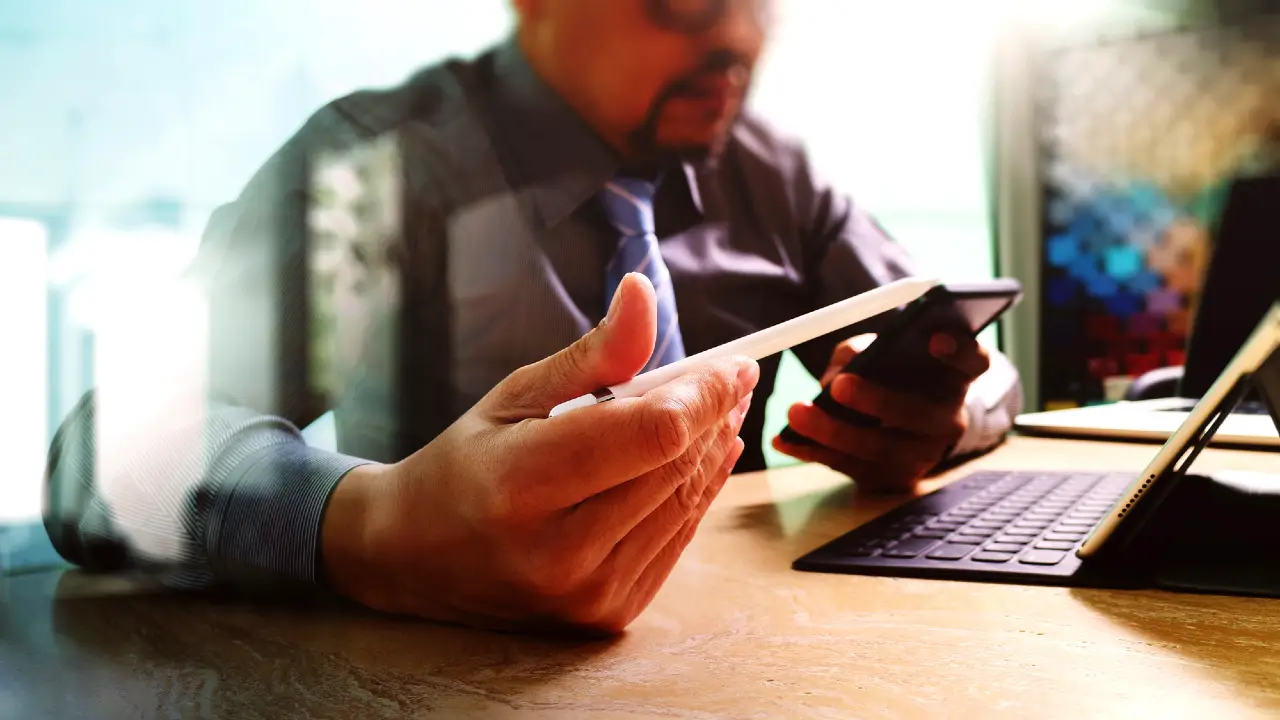 Una persona in camicia e cravatta siede a una scrivania utilizzando un tablet e uno smartphone, con la scritta SEPA cosa significa visualizzata sullo schermo, circondata da una tastiera e da documenti: un ambiente di lavoro intenso.