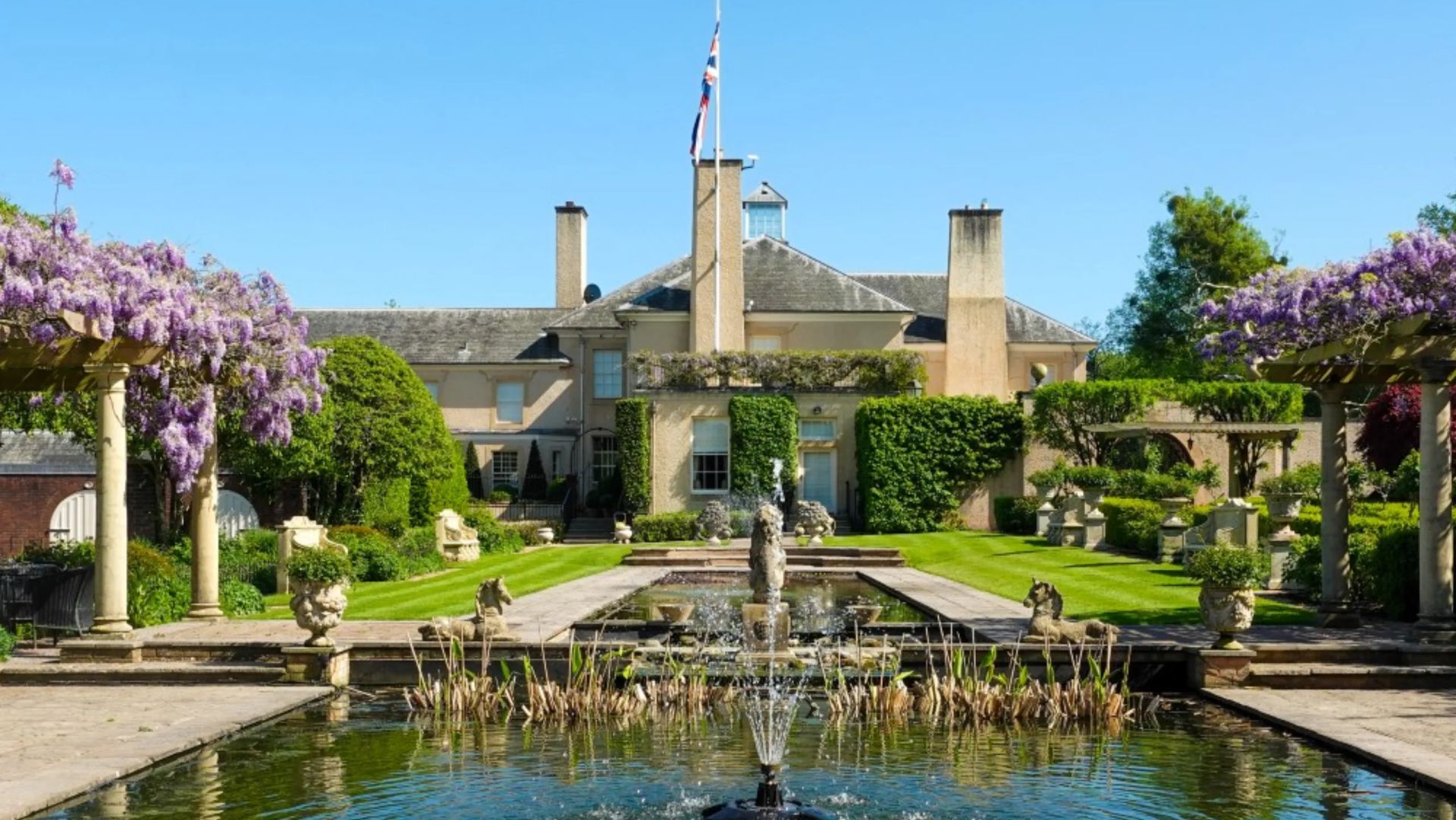 Una grande casa di campagna con muri ricoperti di edera, un pennone centrale, prati curati, cespugli topiati, statue e un laghetto rettangolare con una fontana e pergolati fioriti, il tutto sotto un cielo azzurro e limpido.