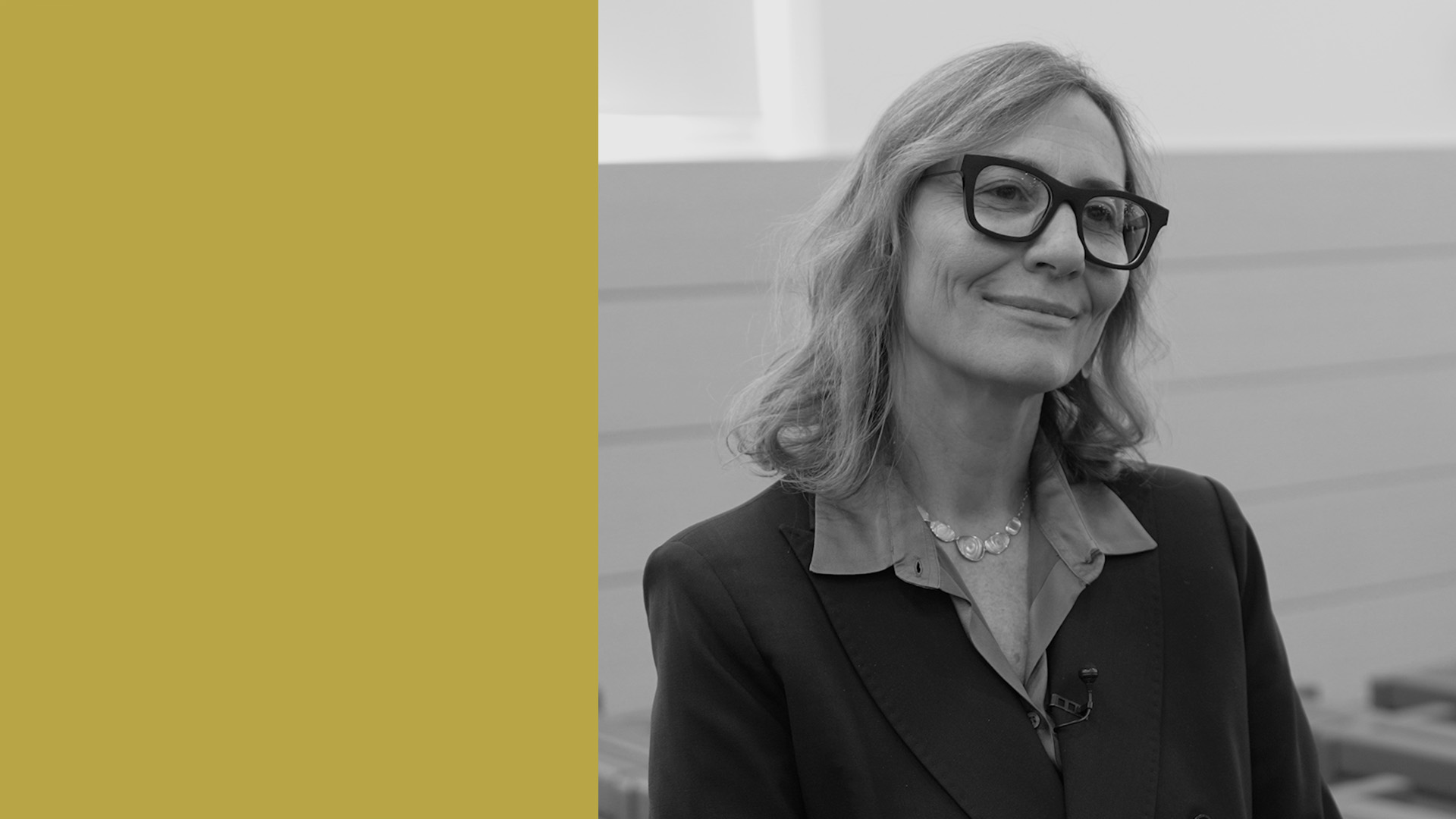 Una donna con occhiali e capelli ondulati, che indossa un blazer e una collana, sorride leggermente mentre si trova in casa. Il lato sinistro dell'immagine è caratterizzato da un blocco solido giallo senape.