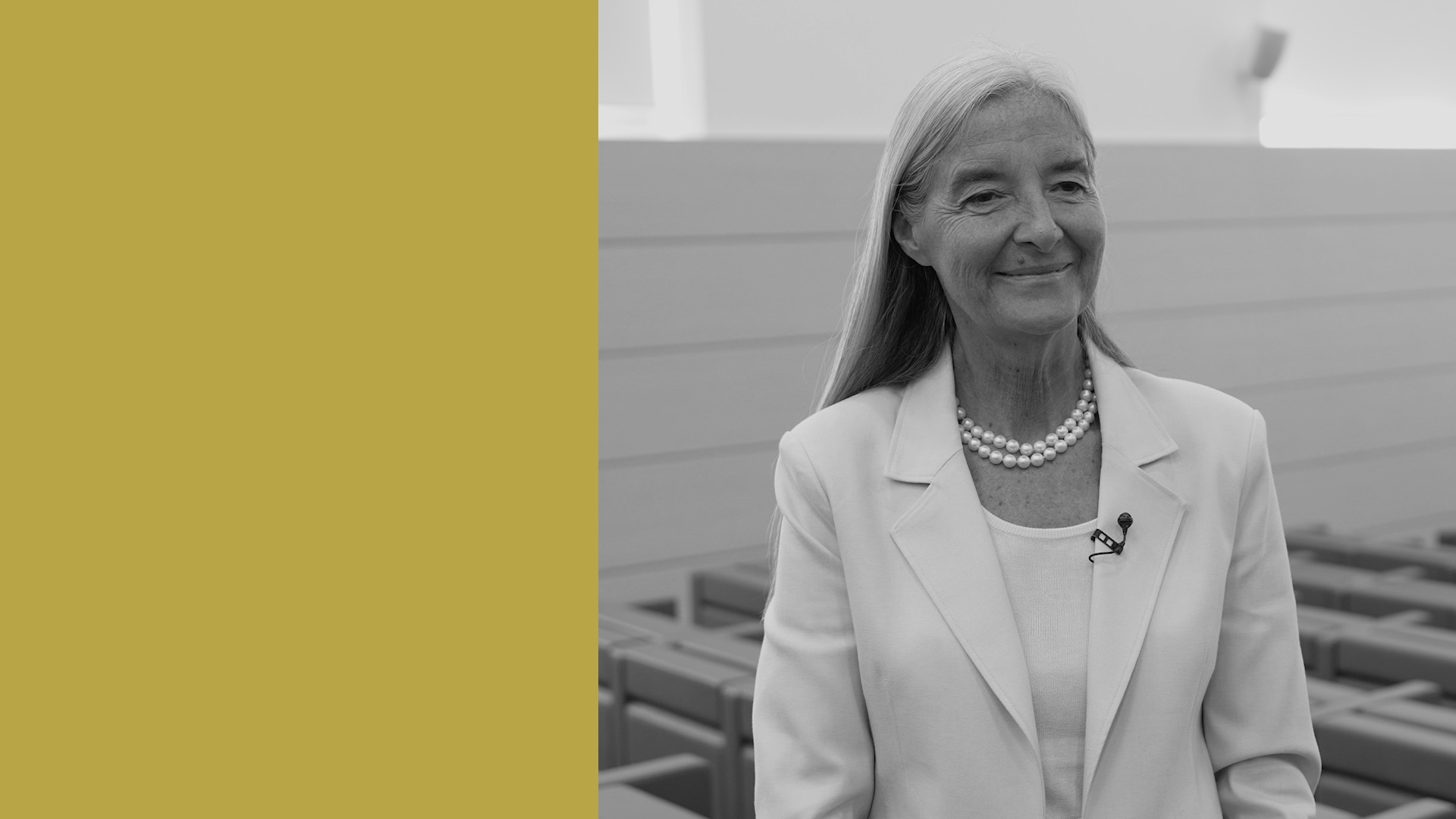 Una donna sorridente con lunghi capelli grigi, che indossa un blazer bianco, una collana di perle e un microfono, si trova in una moderna sala conferenze. Il lato sinistro dell'immagine è riempito da un colore giallo senape pieno.
