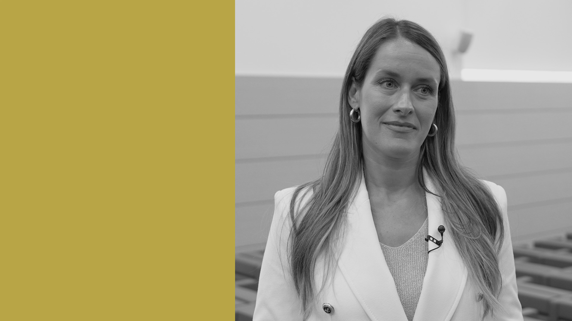 Una donna con i capelli lunghi che indossa un blazer bianco e un microfono a bavero si trova in un luogo chiuso. L'immagine è in bianco e nero con un pannello giallo pieno sul lato sinistro.