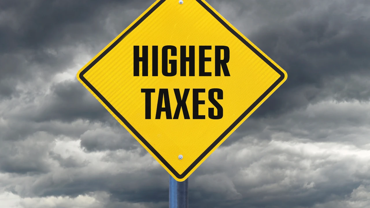 Un cartello stradale giallo a forma di diamante con la scritta "HIGHER TAXES" (tasse più alte) in grassetto nero si staglia contro un cielo scuro e nuvoloso, evidenziando le preoccupazioni per l’aumento della flat tax da 200mila a 300mila euro per Hnwi e grandi patrimoni.