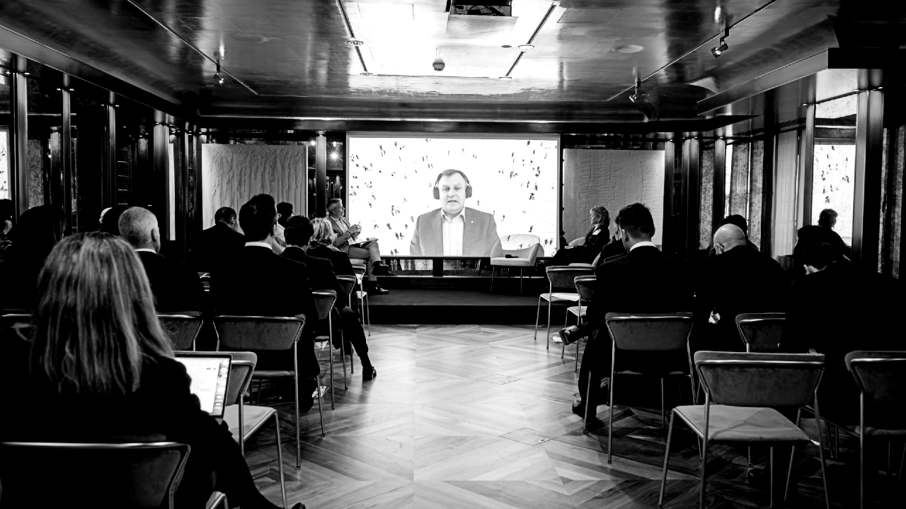 Black and white photo of people seated in a conference room, watching a large screen displaying a man speaking in a virtual meeting. The room has reflective walls and a wooden floor.