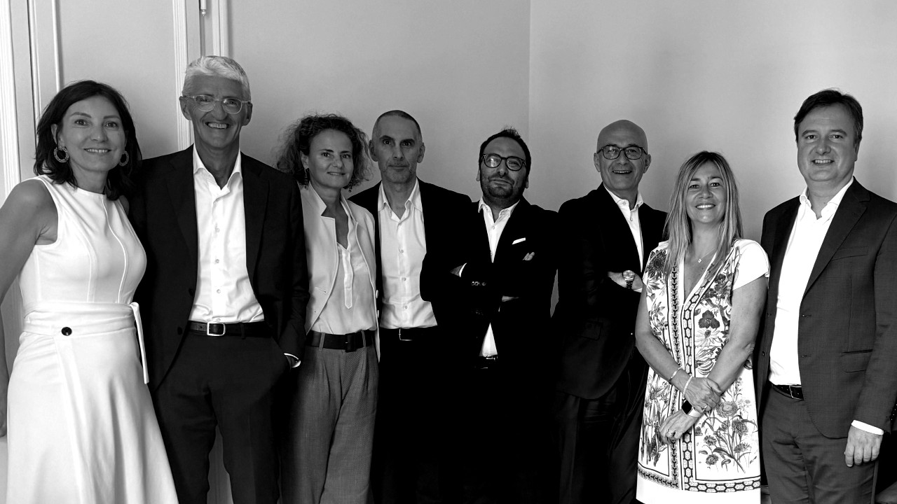 Eight people, dressed in business and smart casual attire, stand side by side and smile for a group photo in a well-lit room with plain walls. The image is in black and white.