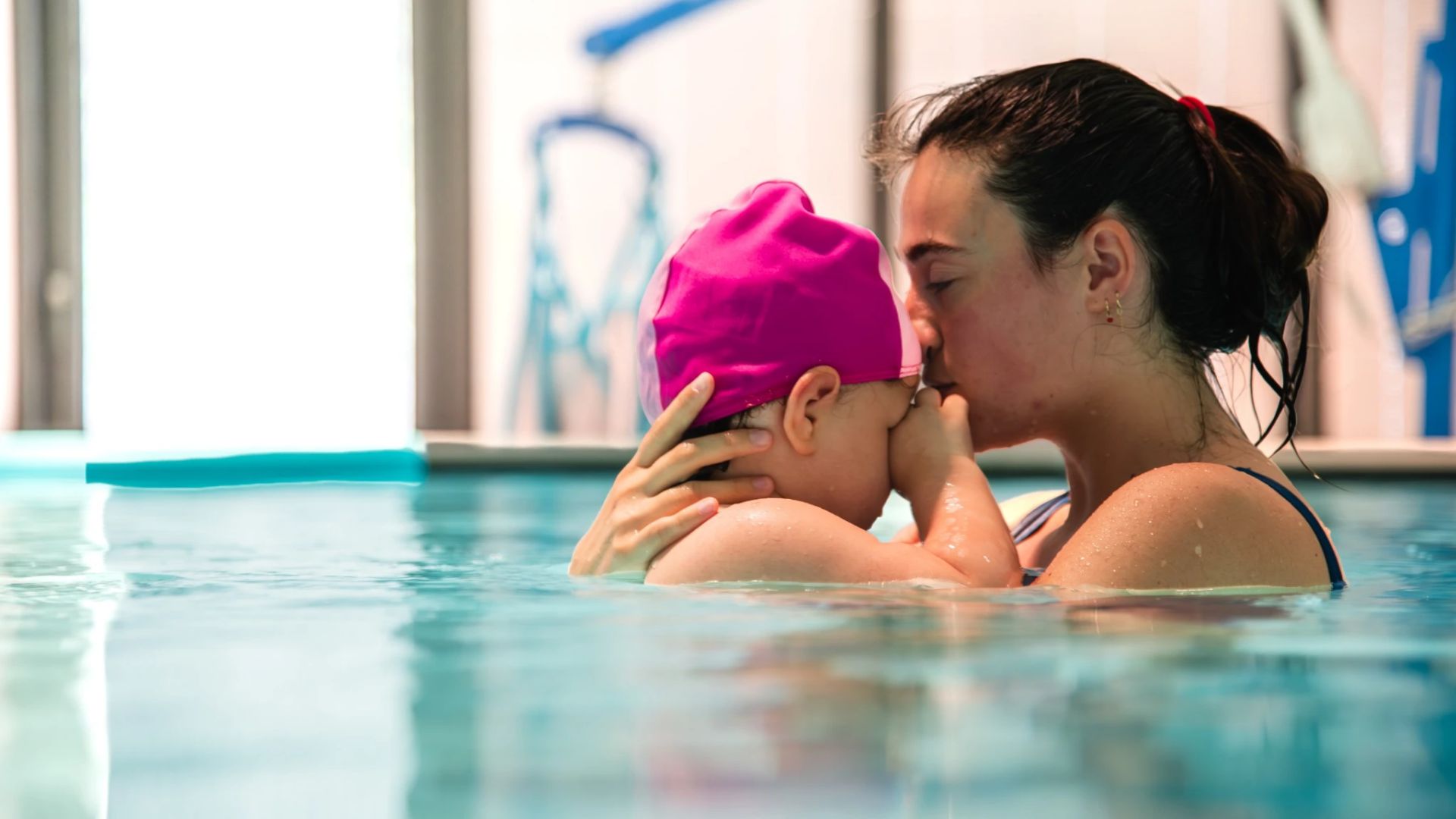 Una donna tiene in braccio e bacia un bambino, che indossa una cuffia rosa, mentre si trovano insieme in una piscina TOG asta. La scena è tenera e affettuosa, con luci soffuse in un'area interna della piscina.