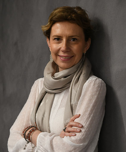 Donna con capelli castano chiaro che indossa una camicetta bianca, un foulard grigio chiaro e braccialetti di perline, in piedi con le braccia incrociate e sorridente davanti a uno sfondo grigio, che rappresenta il viaggio dal private equity alla beneficenza.