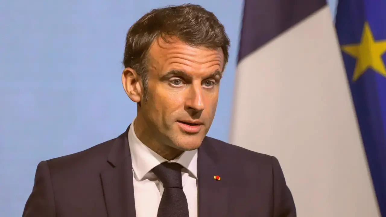 Un uomo in abito scuro e cravatta parla davanti a bandiere francesi e dell'Unione Europea sfocate su uno sfondo azzurro.