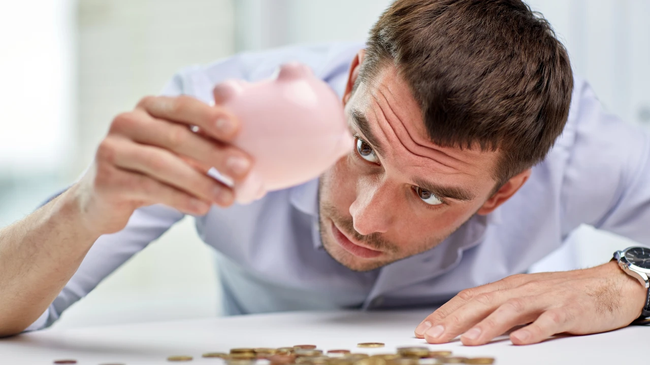 Un uomo inclina un salvadanaio rosa a testa in giù, guardandolo intensamente mentre le monete sono sparse sul tavolo di fronte a lui, catturando le sfide che i giovani devono affrontare tra risparmio e investimenti.