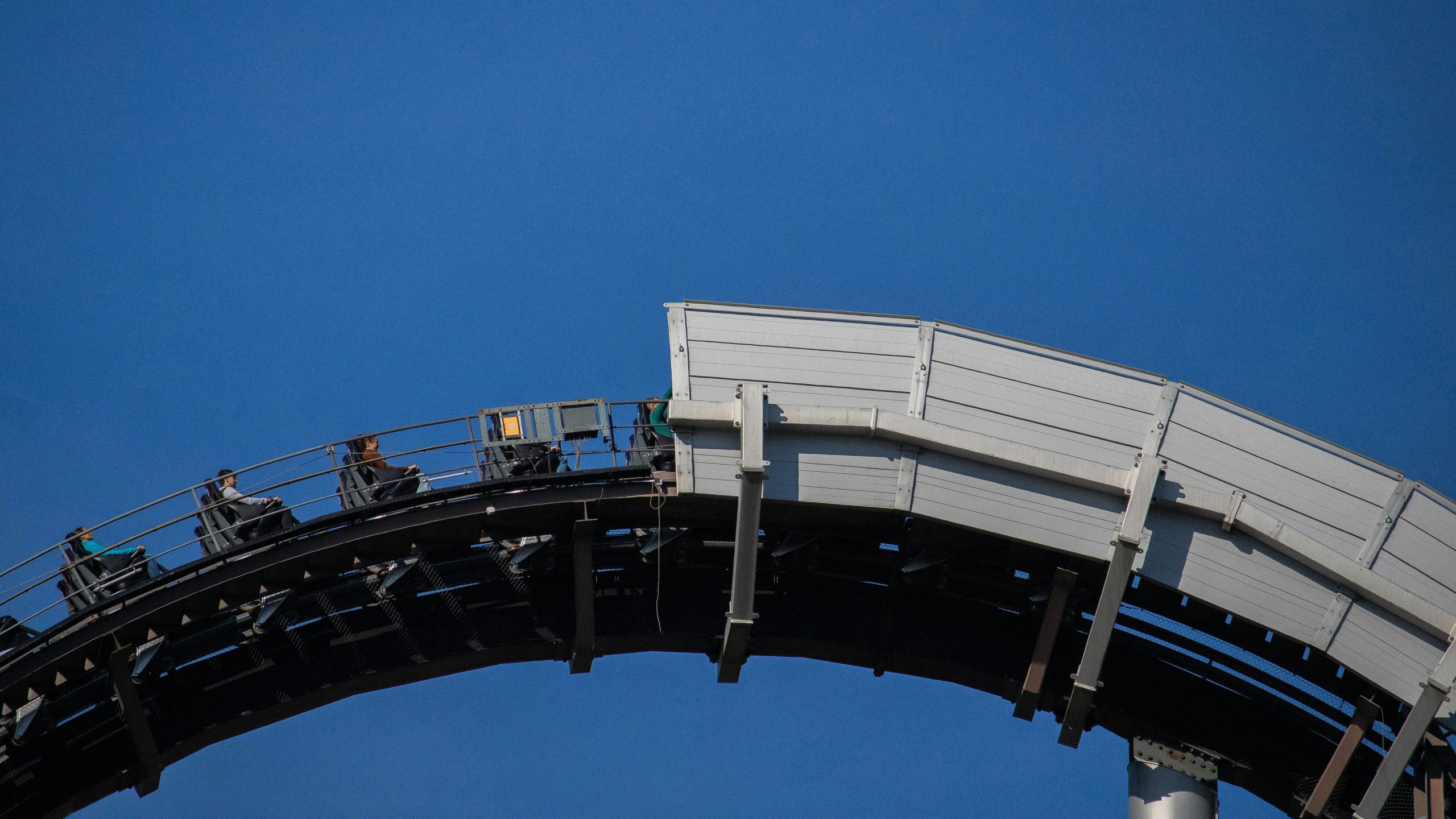 Un ottovolante si arrampica su una pista ripida contro un cielo azzurro e limpido, con alcuni passeggeri visibili vicino alla cima, che si preparano per l'imminente caduta.