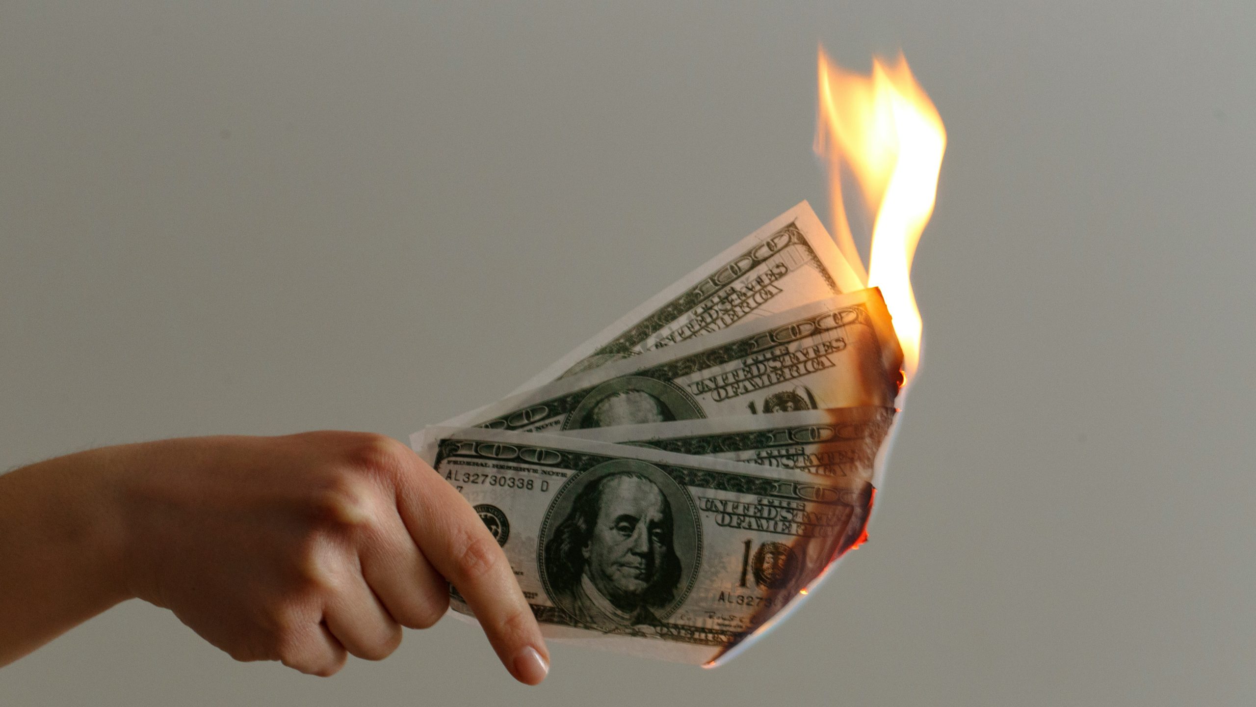 Una mano che regge diverse banconote da cento dollari statunitensi in fiamme, con le fiamme che consumano un angolo, su uno sfondo chiaro.