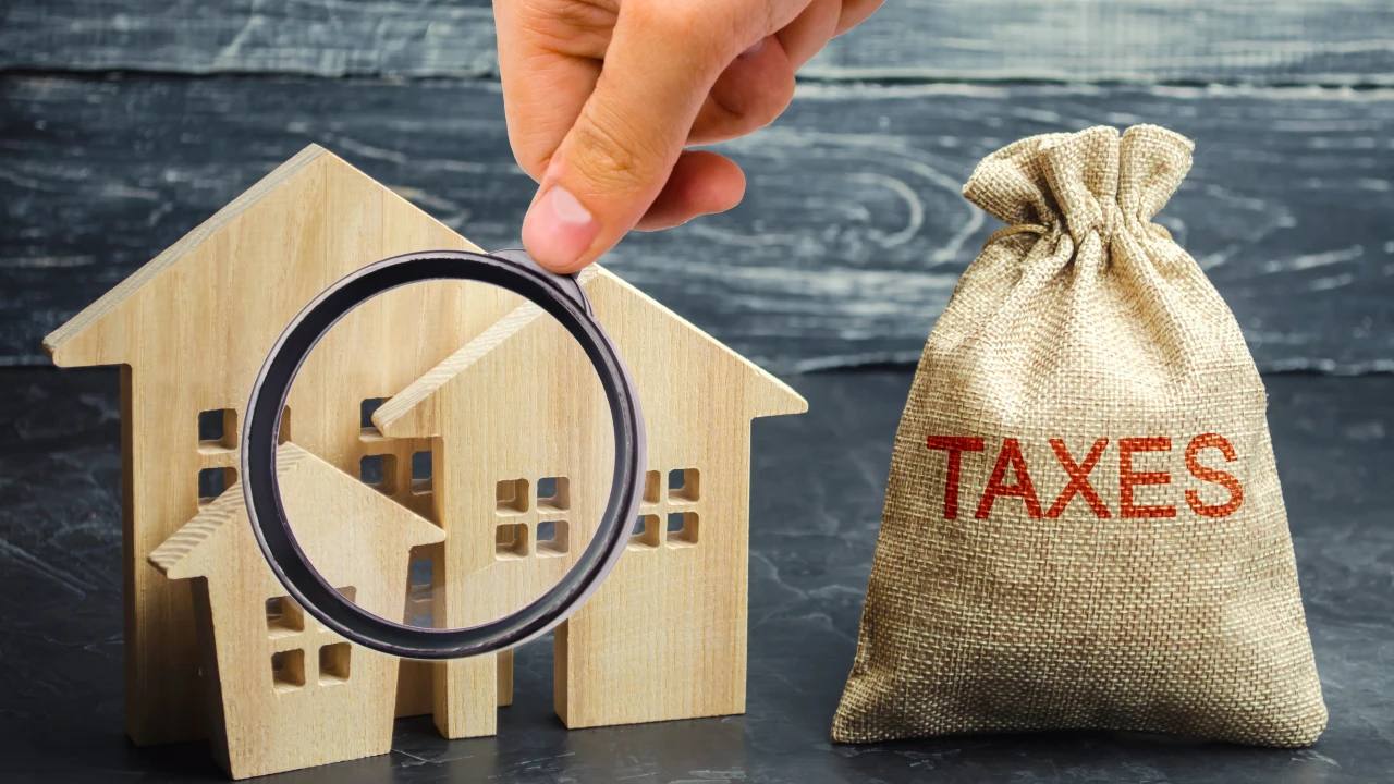 Una mano tiene una lente d'ingrandimento su piccoli modelli di case in legno accanto a un sacco di iuta con la scritta "TAXES", che simboleggia l'accertamento delle imposte sulla proprietà o le tasse immobiliari. Il riferimento va alla stangata della Manovra 2026 sugli affitti brevi, con innalzamento della cedolare secca dal 21 al 26%.