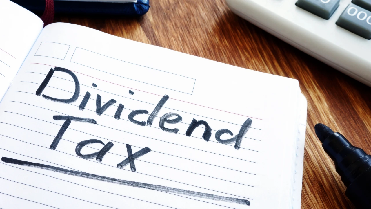 Un quaderno con la scritta "Dividend Tax" scritta con pennarello nero su una pagina foderata, posto su una scrivania di legno accanto a una calcolatrice e a una penna. Il riferimento va alla stretta della manovra 2026 sui dividendi.