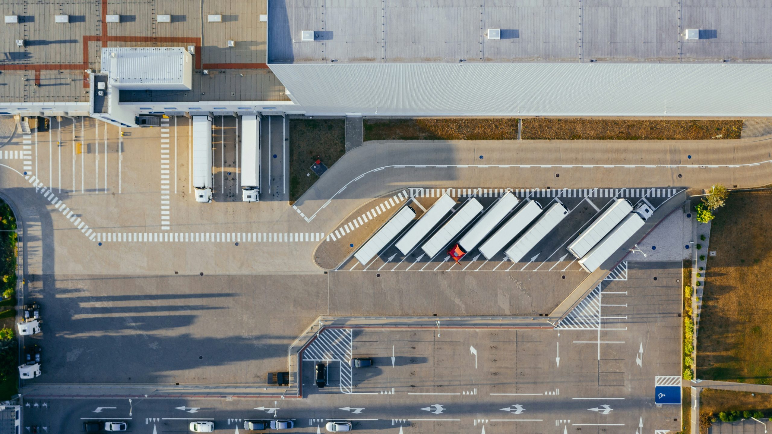 Vista aerea di una banchina di carico di un magazzino con diversi semirimorchi bianchi parcheggiati nelle baie, tetto grigio dell'edificio e strade segnalate con strisce pedonali e veicoli nell'area circostante.