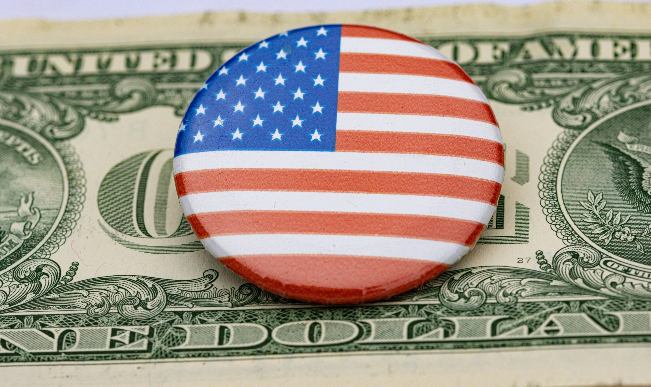 Una spilla rotonda con il disegno della bandiera americana poggia su una banconota statunitense da un dollaro, coprendo parzialmente la parola "ONE" e parte di "UNITED STATES OF AMERICA".