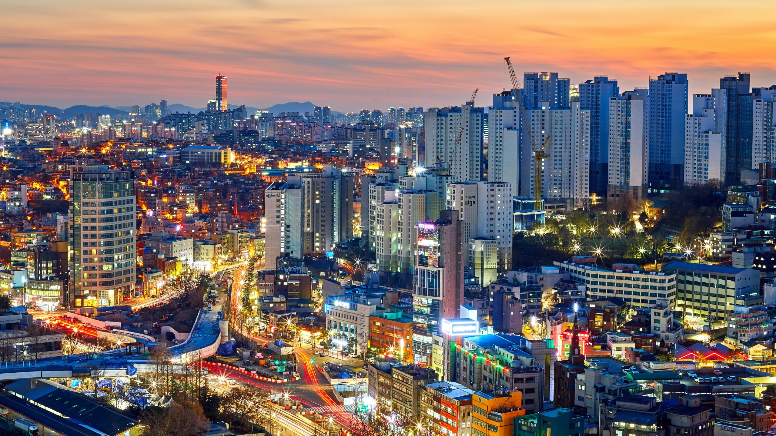 Un vibrante paesaggio urbano al tramonto con alti grattacieli, edifici illuminati e strade affollate di traffico in movimento, che creano una vivace atmosfera urbana su un cielo arancione e blu.